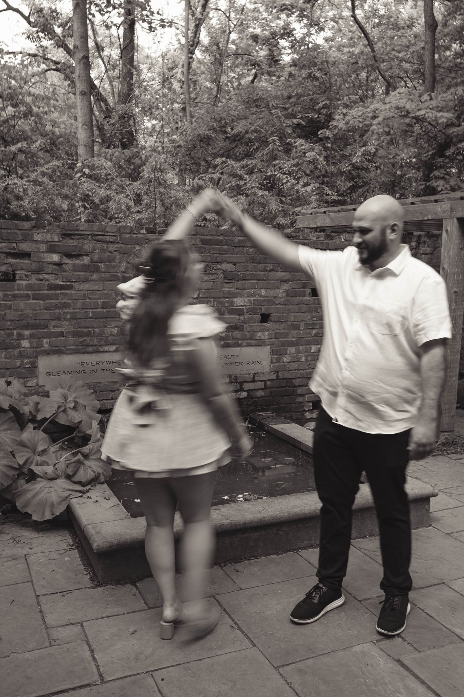 A bald bearded man twirls a woman in a sundress while dancing in a brick courtyard surrounded by trees.