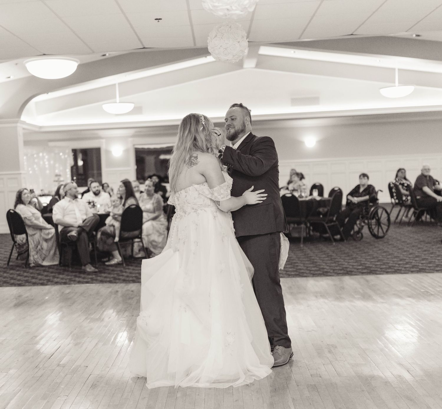 Bride and groom share first dance in ballroom while wedding guests watch in black and white photo.