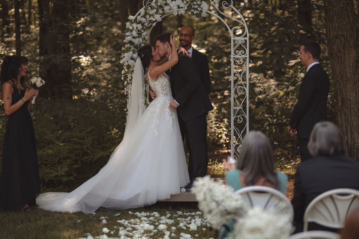 Bride and groom share first kiss under floral arch at outdoor woodland wedding ceremony.