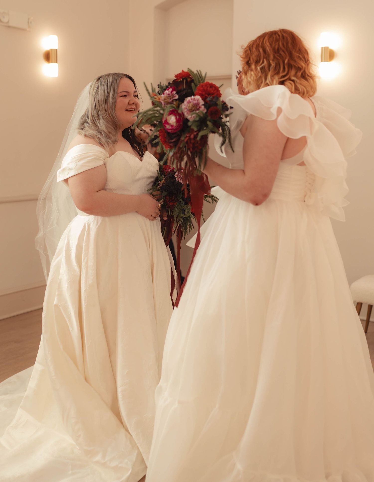 Two brides in elegant white wedding dresses share a joyful moment with a lush bridal bouquet.
