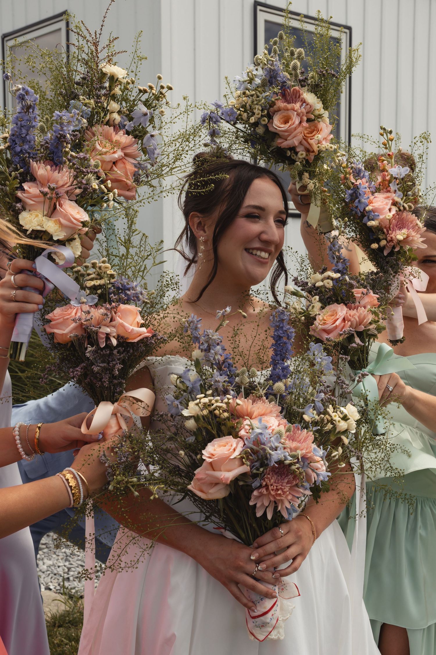 Smiling bride in white dress holds pink rose bouquet, framed by floral wreath with pink dahlias and purple flowers outdoors.