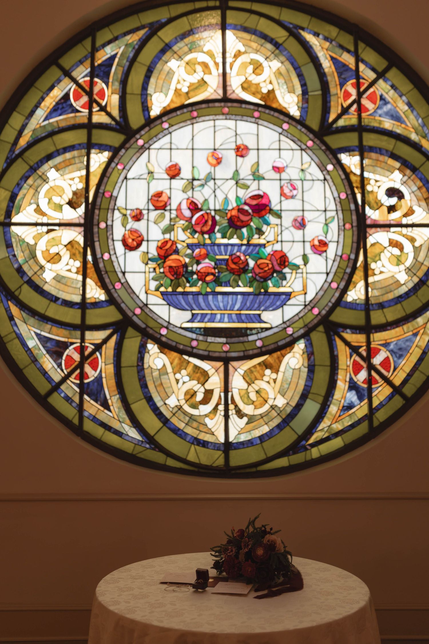 Ornate circular stained glass window featuring roses in a vase, above a small table with flowers.