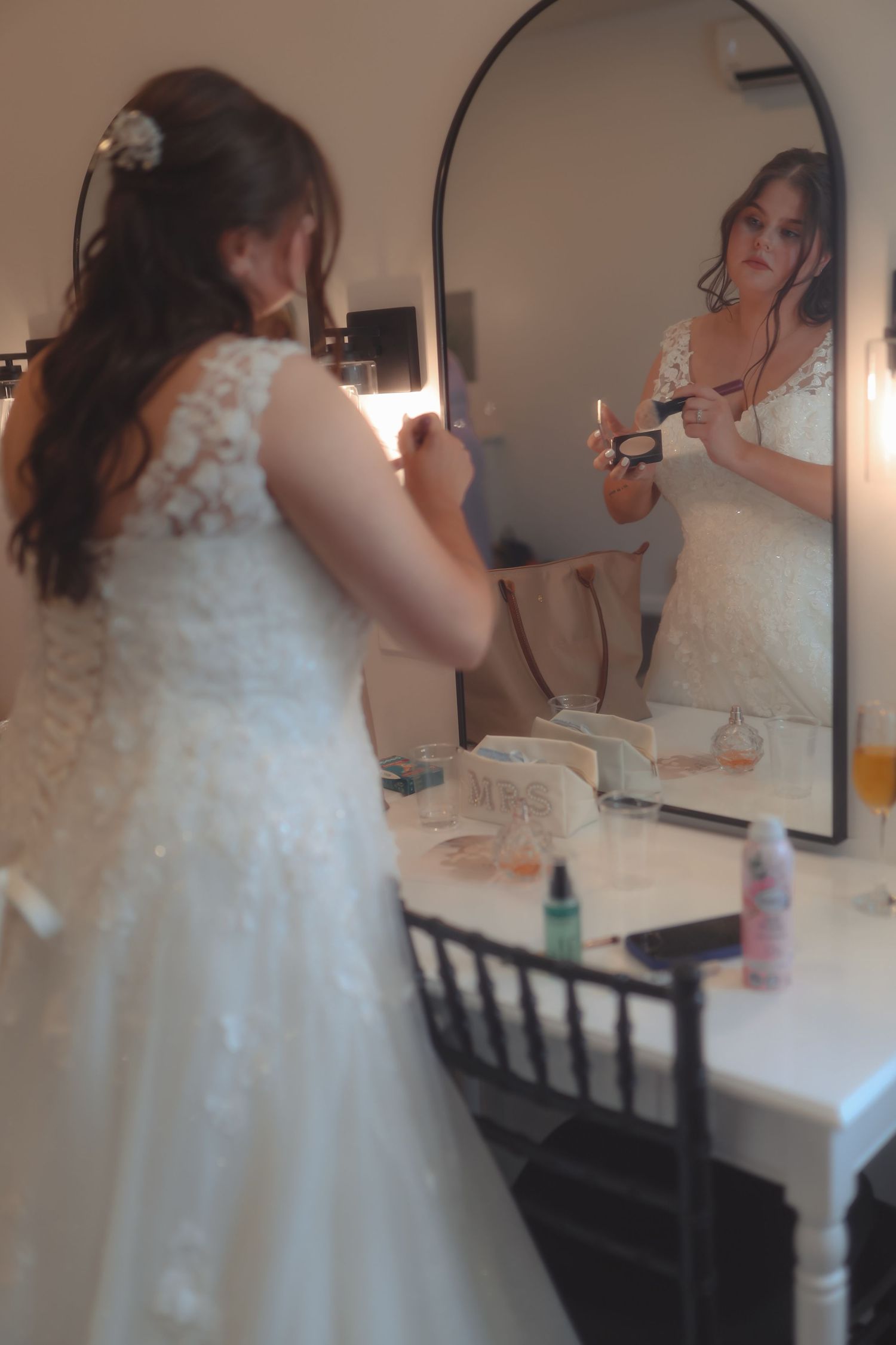 Bride in white lace gown applies makeup at vanity mirror while getting ready for her wedding day.