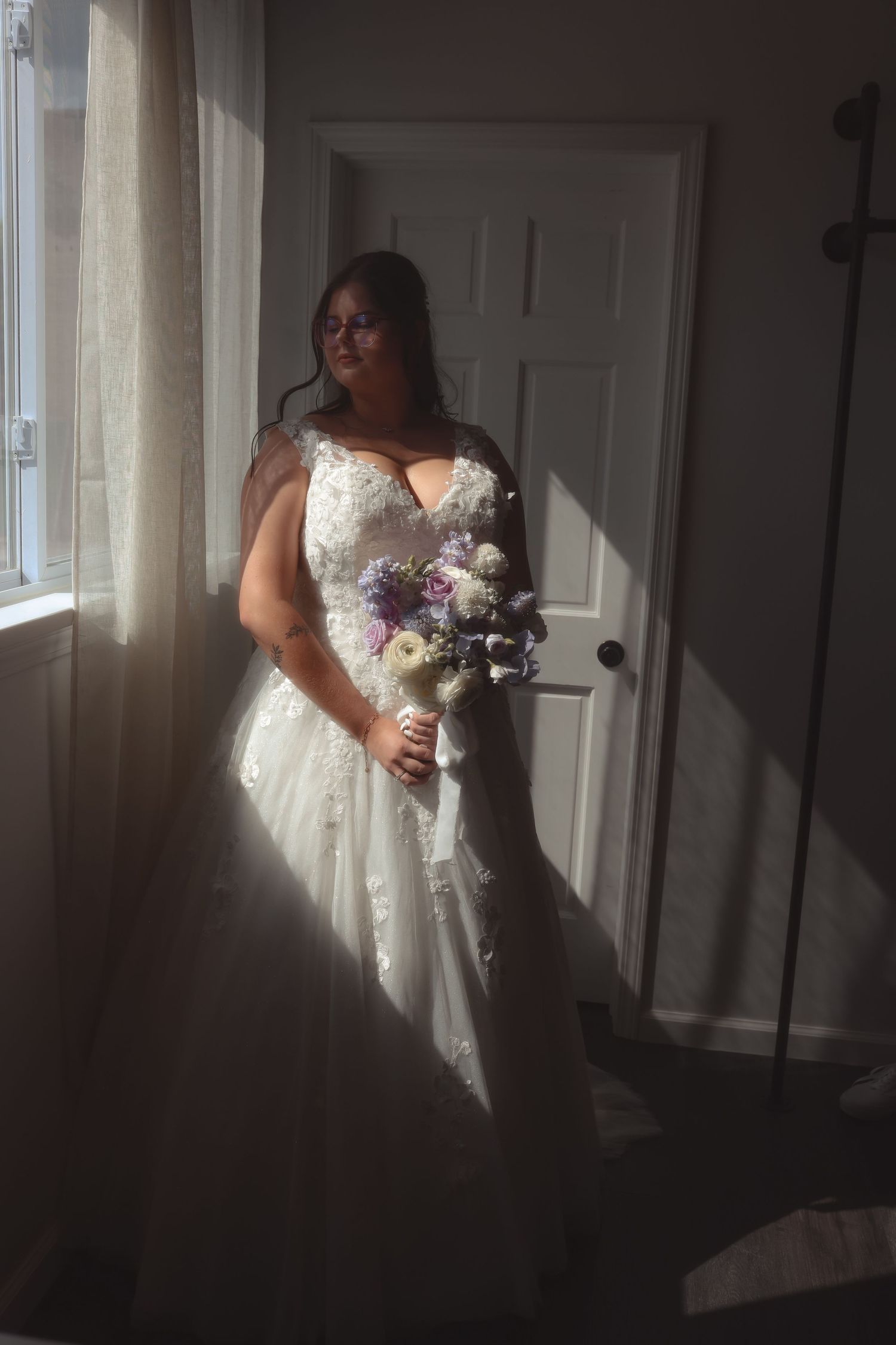Bride in white lace wedding gown holding purple and white bouquet standing by sunlit window.