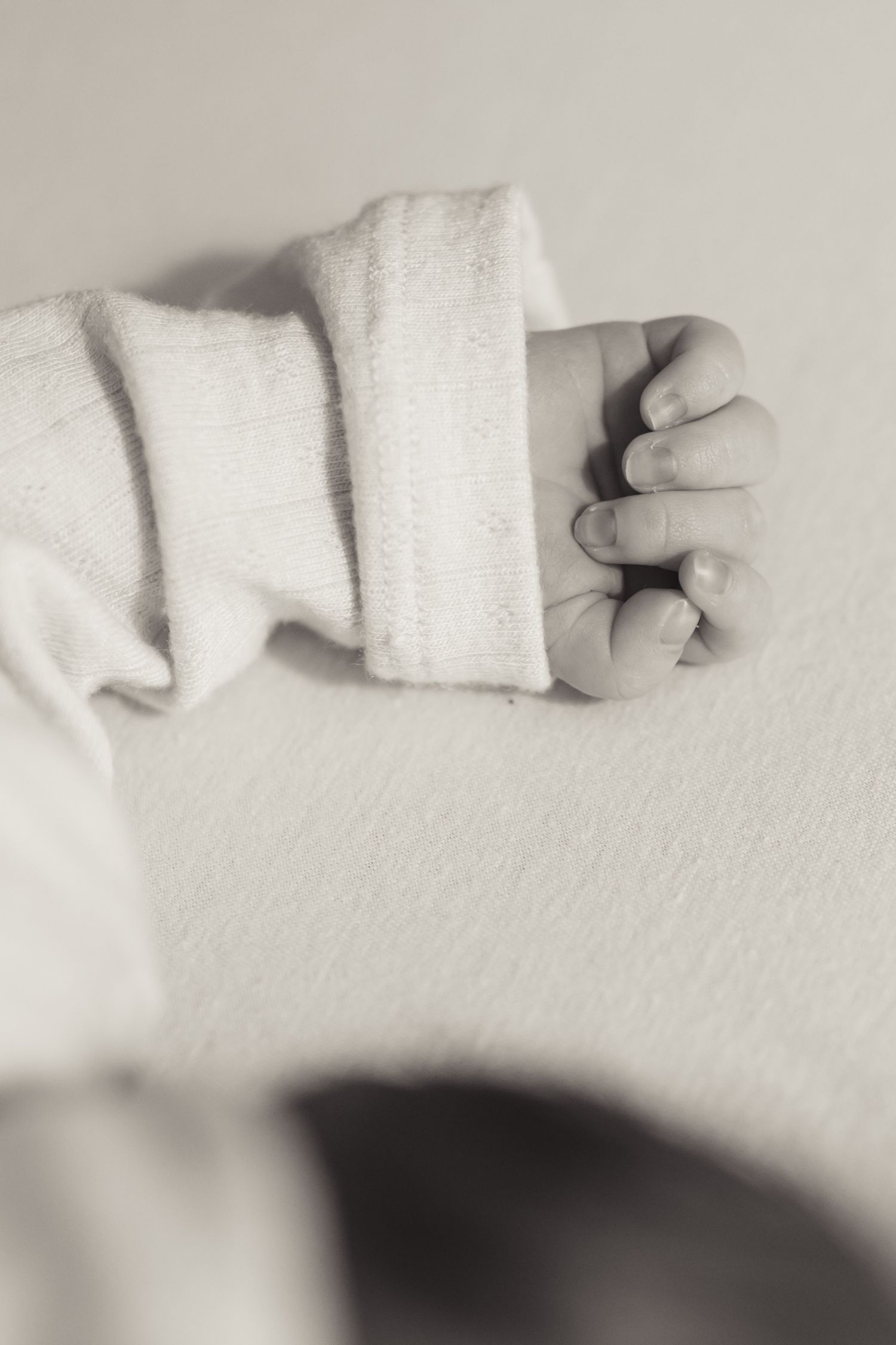 Close-up black and white photo of a newborn baby's tiny clenched fist in a white onesie.