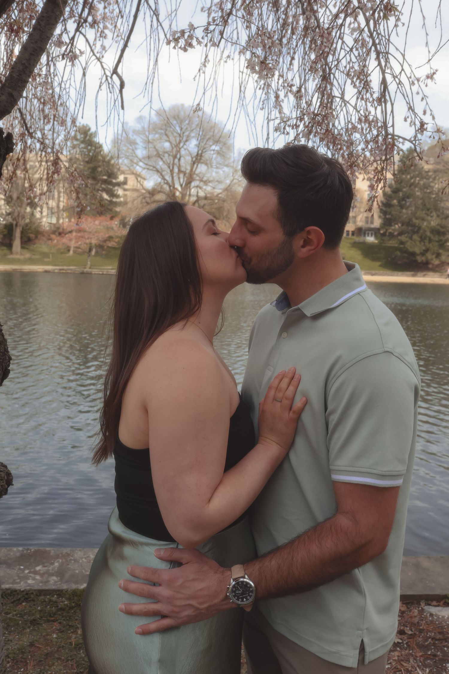 Couple sharing a romantic kiss by a serene lake, surrounded by bare trees in a lush park setting.
