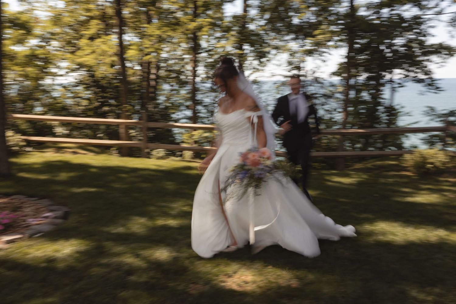 Bride in white gown holding bouquet walks with groom in suit across green lawn at outdoor wedding ceremony.