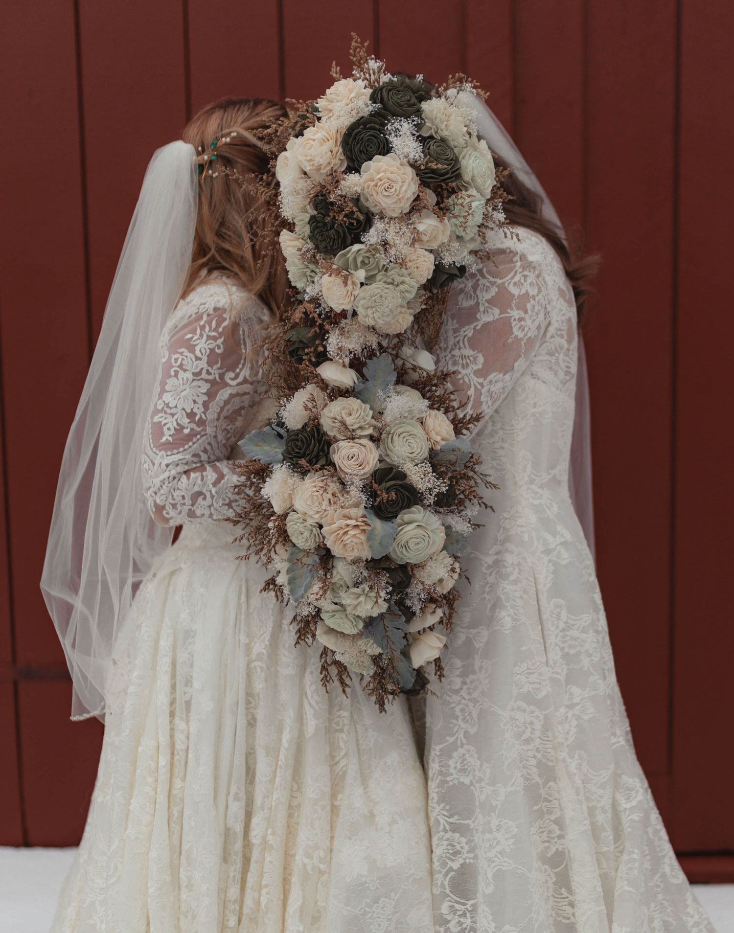 Two brides in lace wedding dresses holding an elegant cascading bouquet of cream and sage flowers against a red barn.