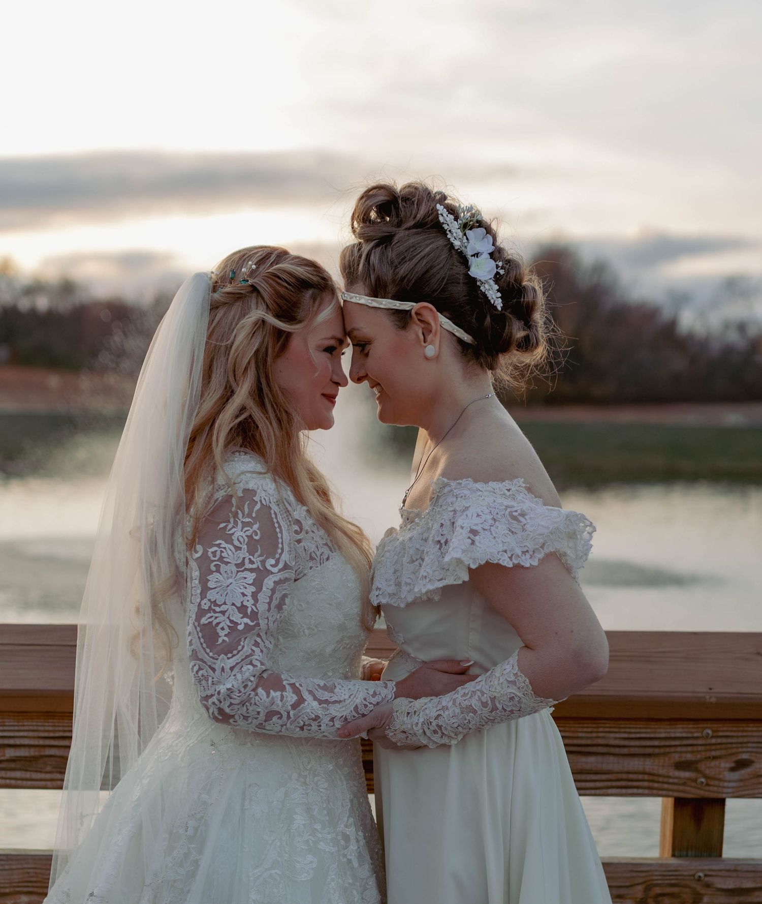 Two brides in white lace wedding gowns share an intimate moment on a wooden bridge at sunset.