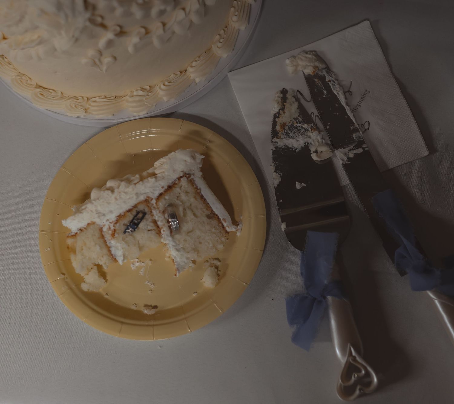 A slice of white wedding cake on a yellow plate beside cake serving utensils with blue ribbon handles.