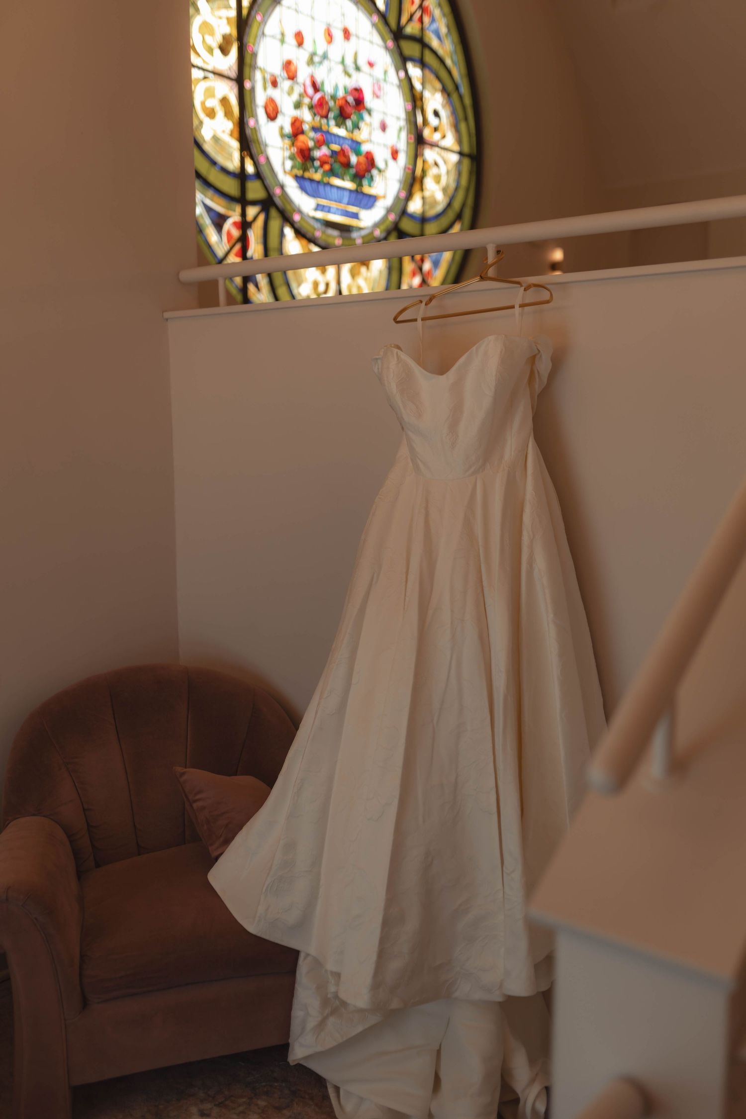 White wedding gown hanging on a hanger in front of a colorful stained glass window in a church.