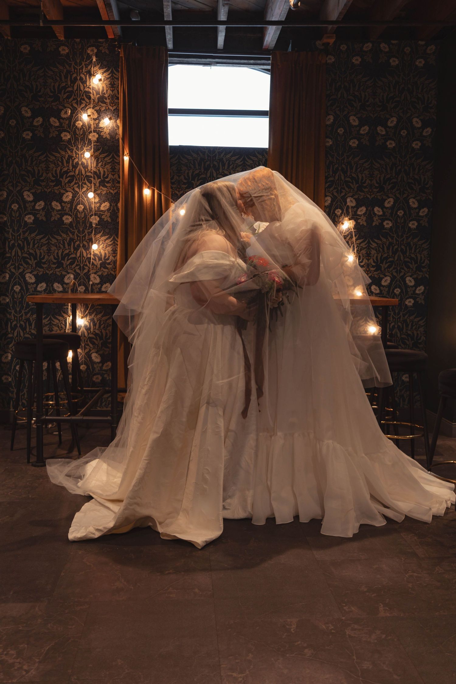 Two brides in white gowns share a kiss under a flowing veil in a warmly lit, decorated venue.