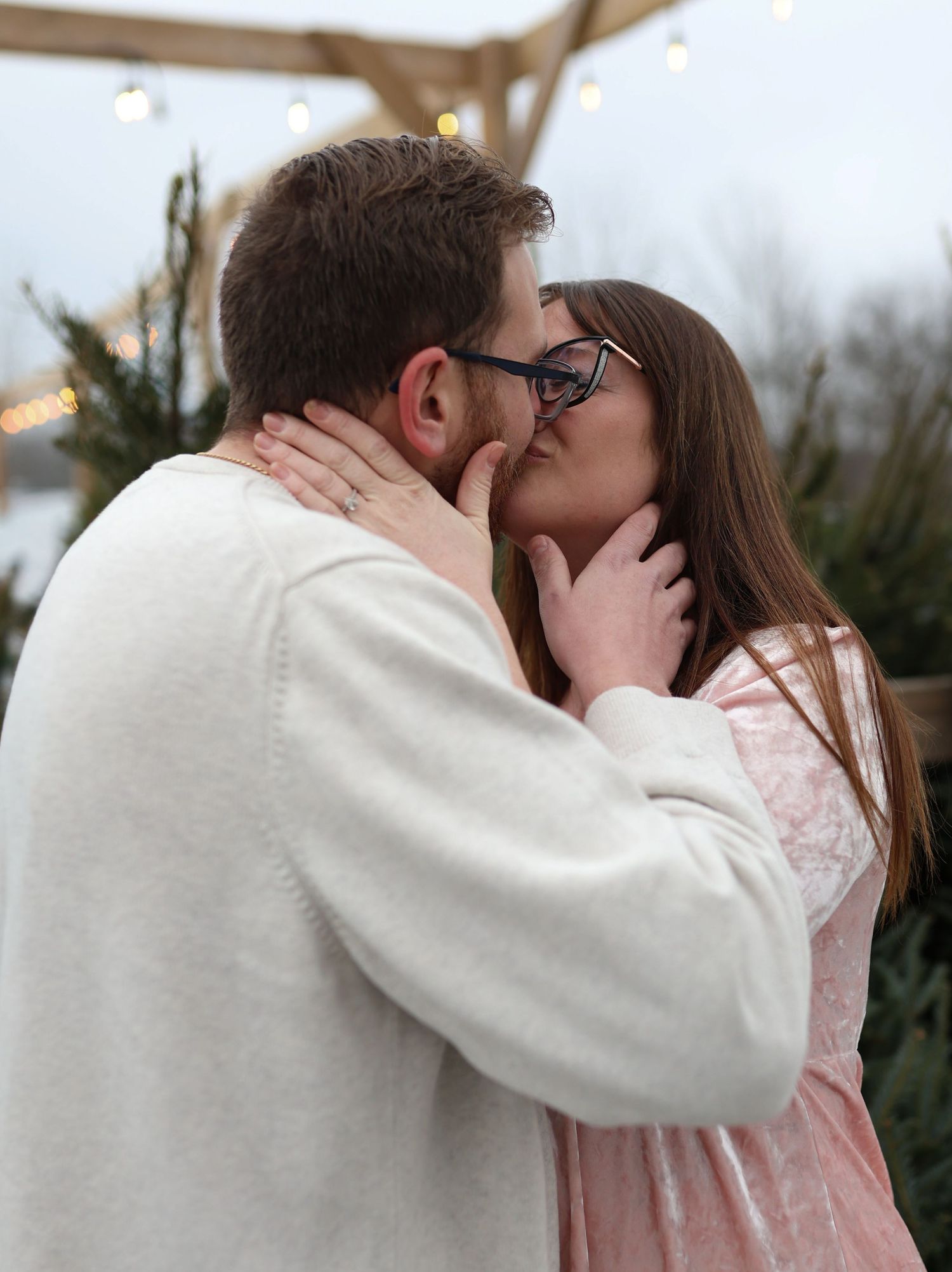 Couple sharing a romantic kiss outdoors at a Christmas tree farm with string lights in the background.