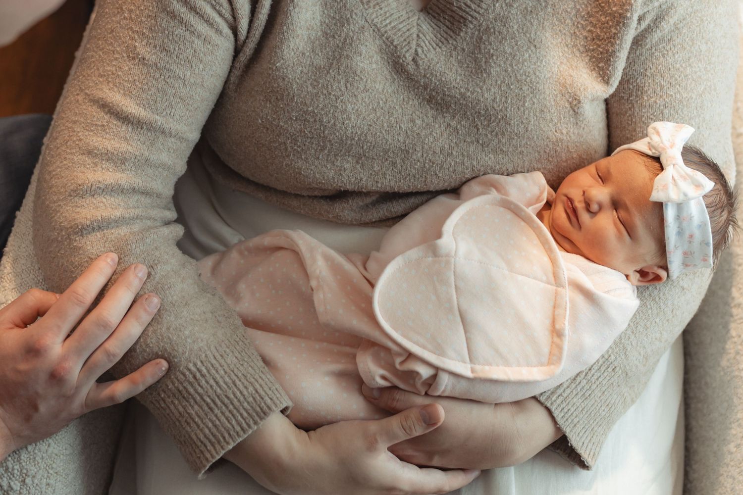 Newborn baby swaddled in white blanket with bow headband, sleeping peacefully in parent's arms.