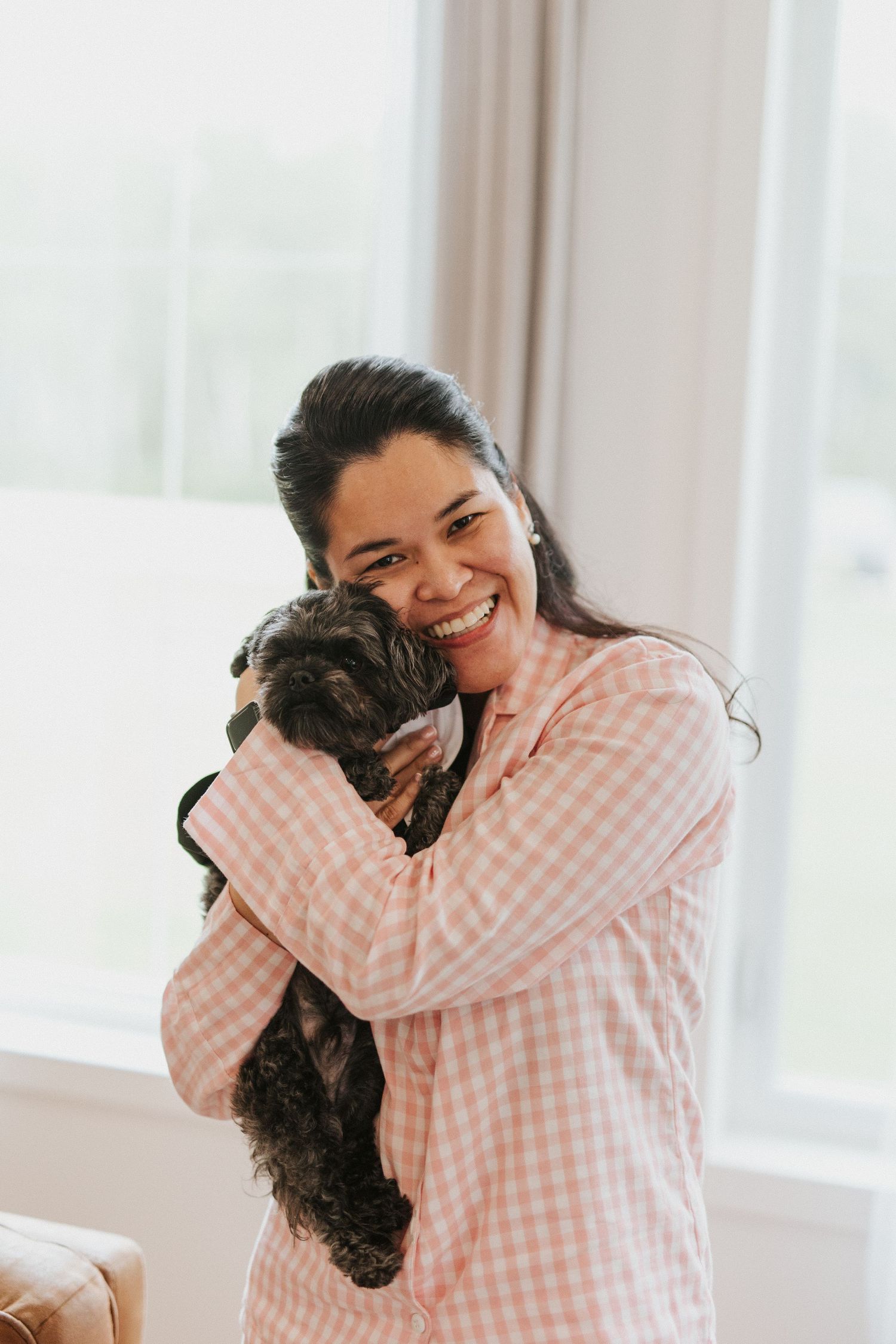 Someone wearing a pink gingham shirt hugs and smiles with their small black dog by a bright window.