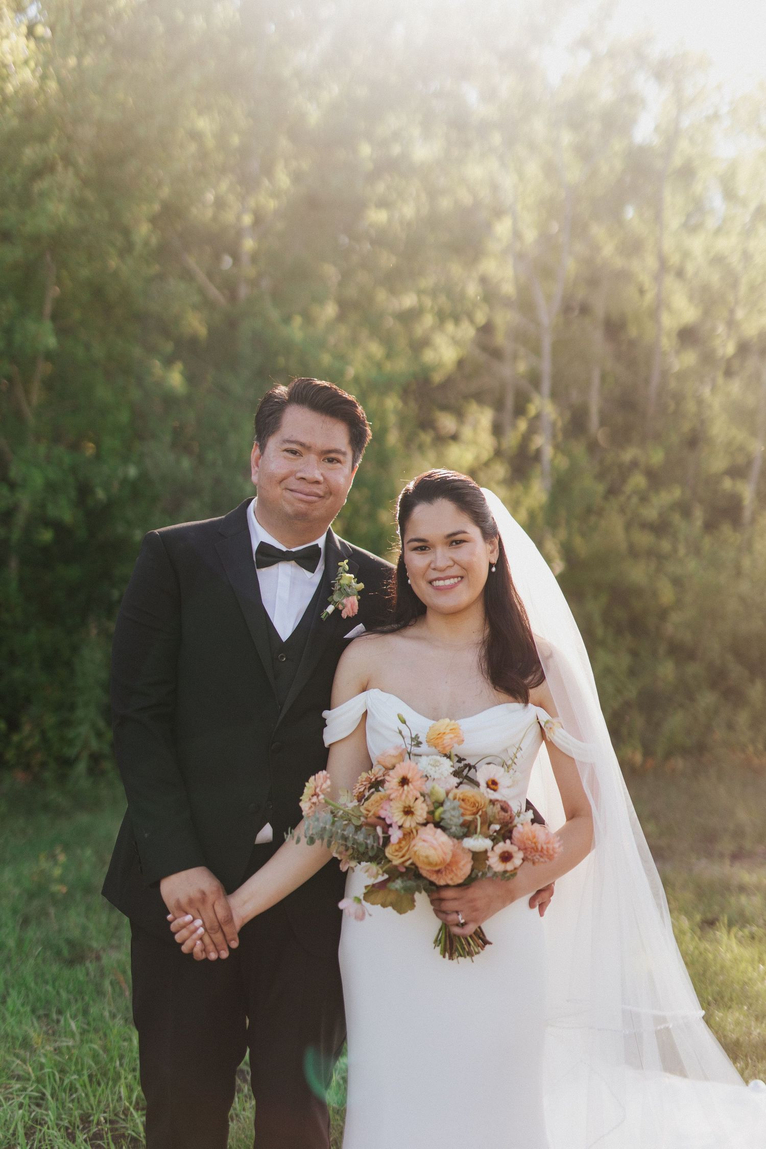 A happy wedding couple poses together in a sunlit outdoor setting with a peach and white floral bouquet.