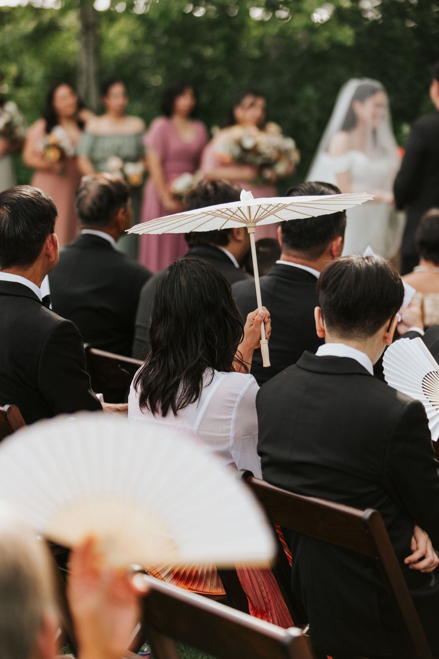 Wedding ceremony guests seated outdoors holding decorative paper parasols during summer celebration.