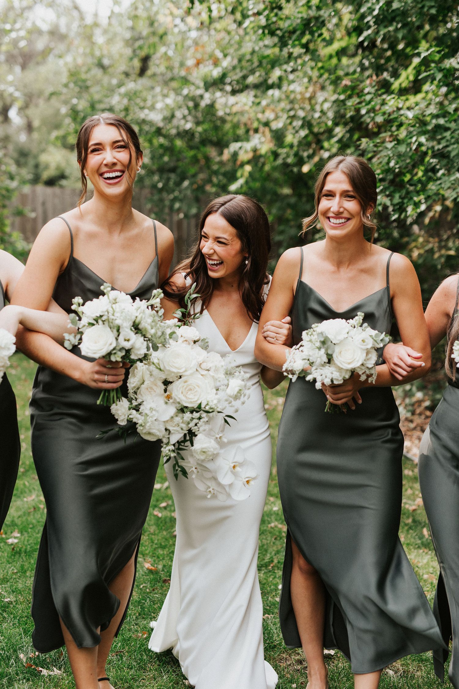 Wedding party in sage green dresses and white gowns holding matching bouquets outdoors.