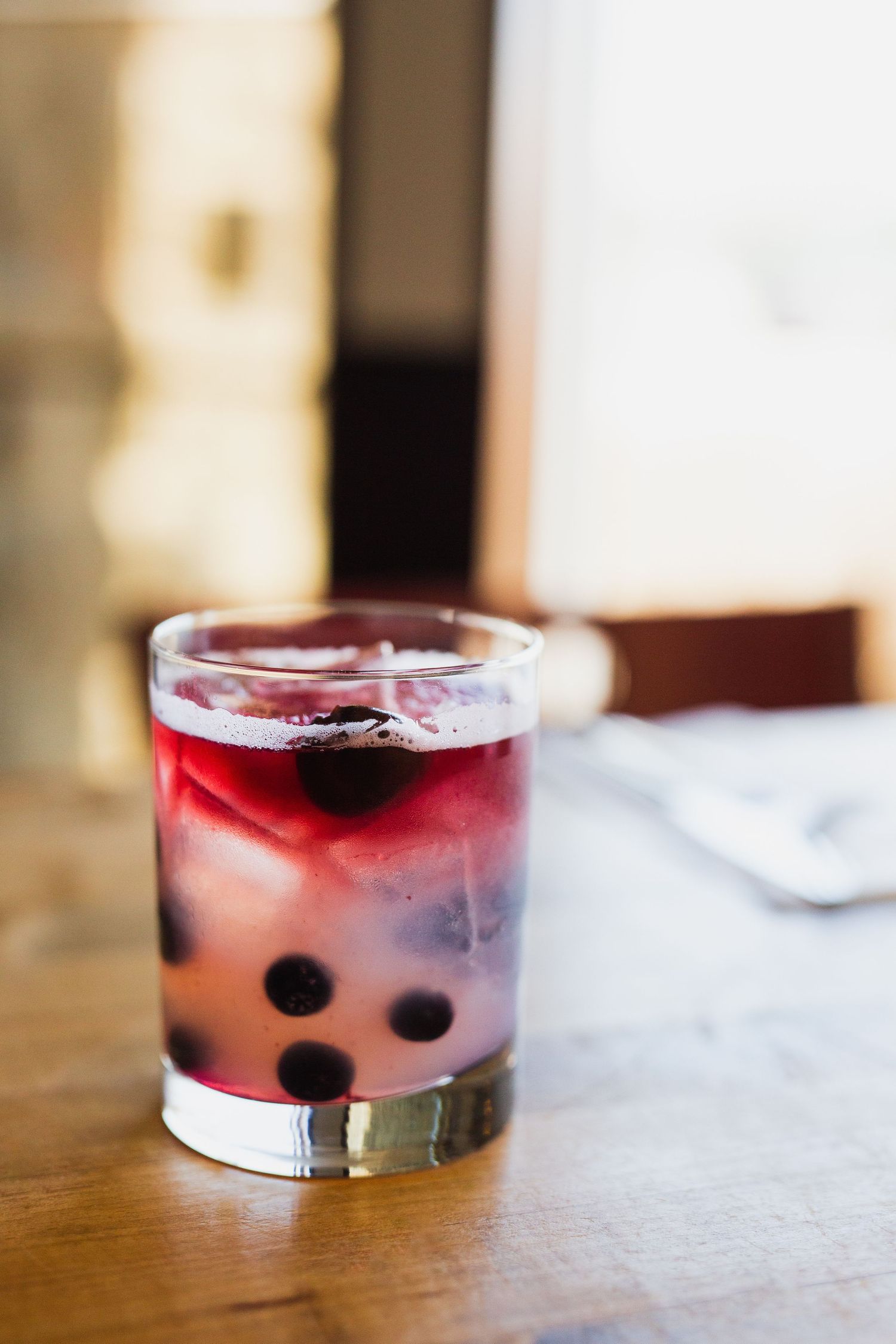 Red cocktail garnished with dark berries in a clear glass on wooden surface.