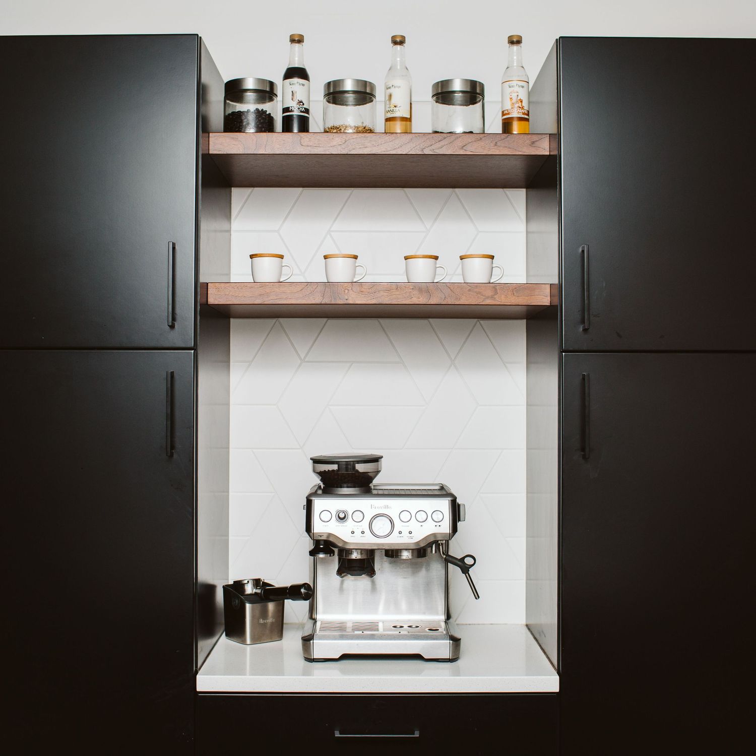 Coffee station with espresso machine and pendant lights mounted on white backsplash with dark cabinets.