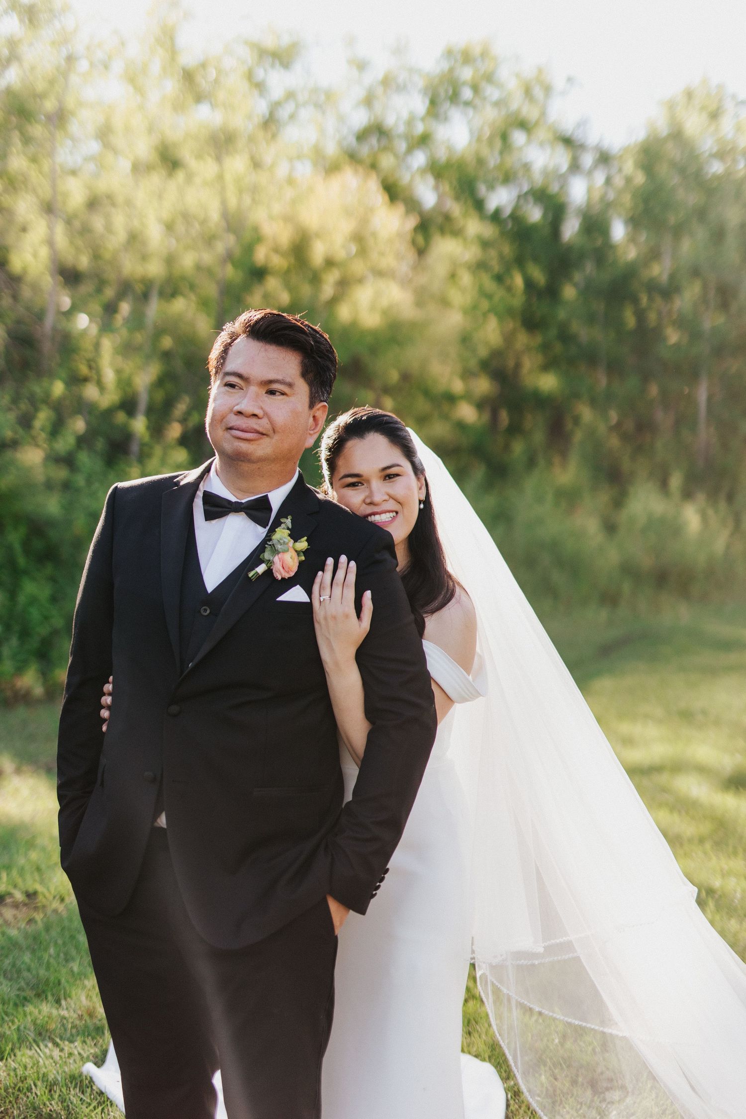 A joyful wedding couple shares an intimate moment outdoors with sunlit trees in the background.