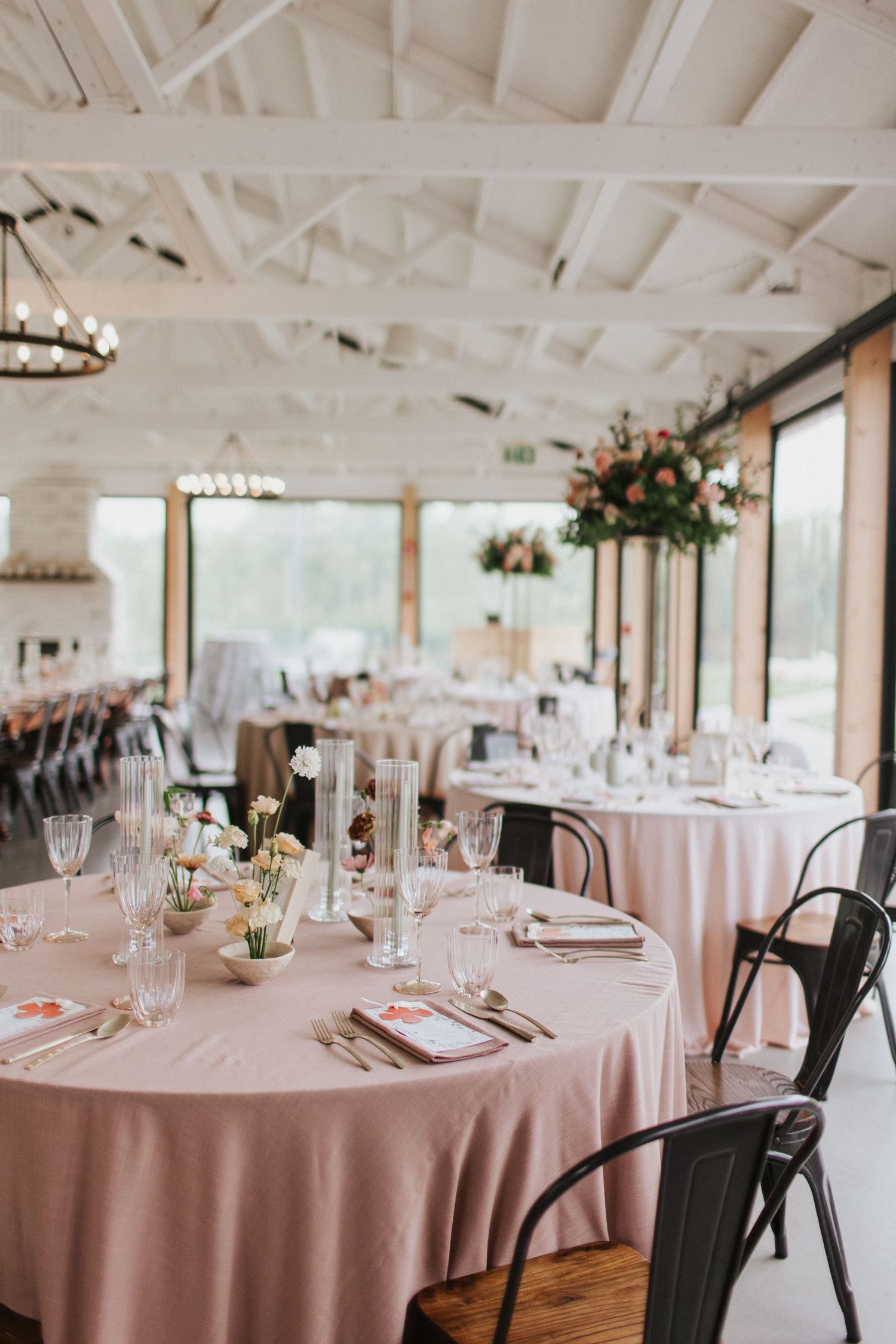 Elegant wedding reception venue with pink tablecloths, industrial chairs, and floral centerpieces under white beamed ceiling.