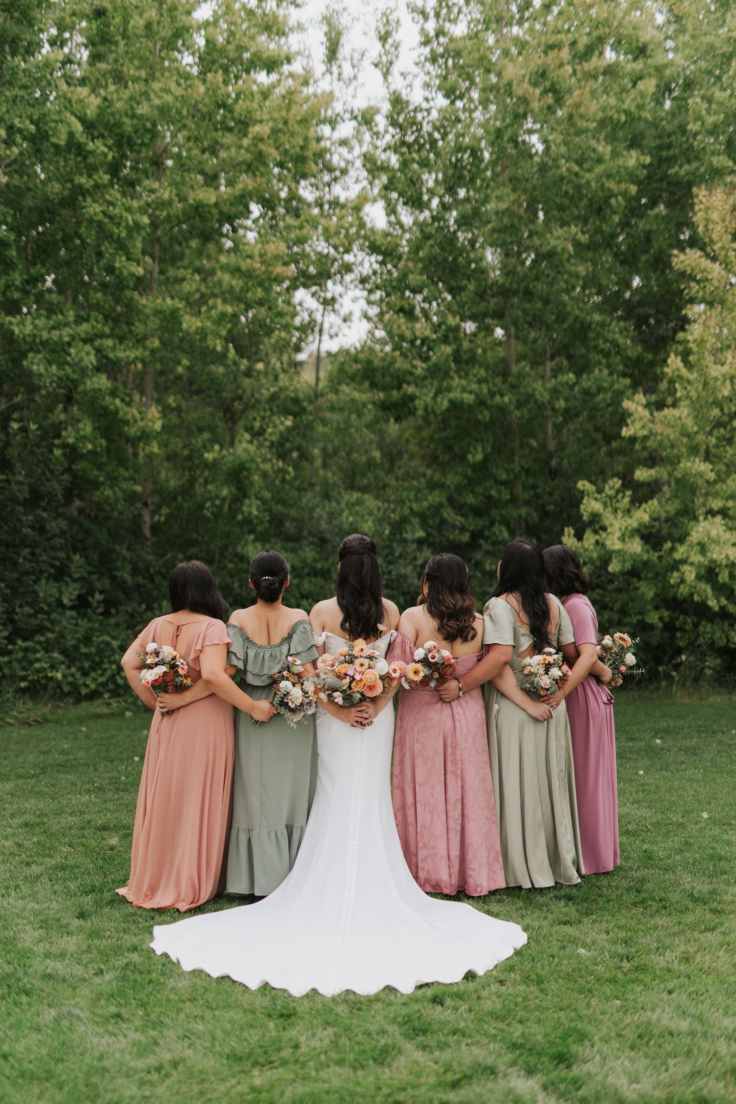 Bridesmaids in blush and sage dresses stand together with the bride in a white gown on a grassy lawn.
