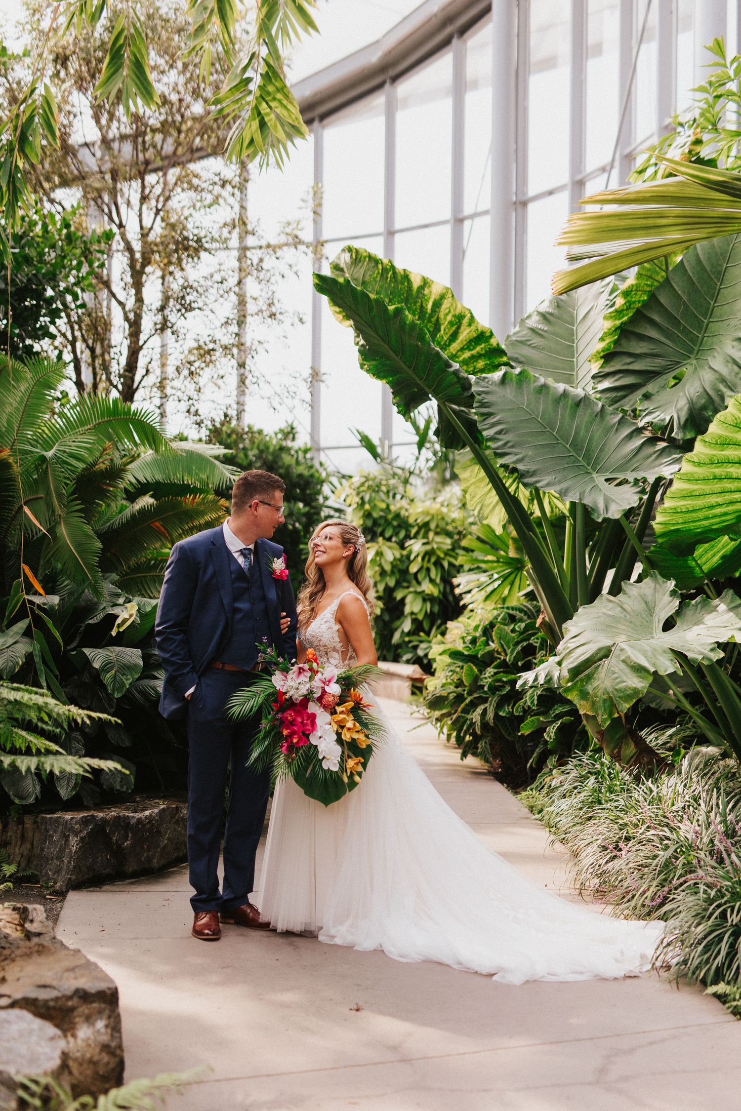 A couple poses in a lush greenhouse conservatory with tropical plants and white stone pathways.