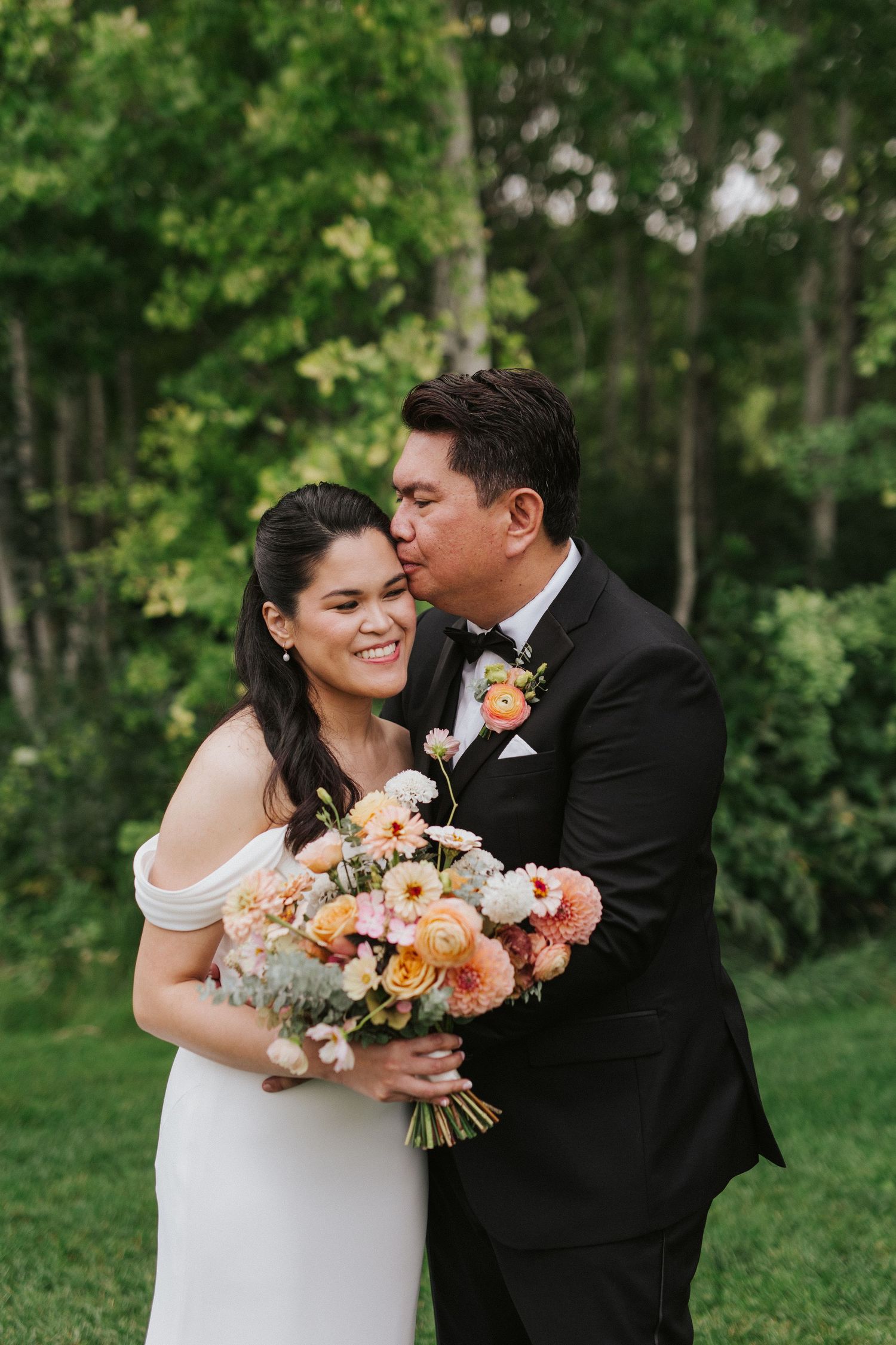 A romantic wedding couple embraces while holding a vibrant peach and coral floral bouquet in a wooded garden.
