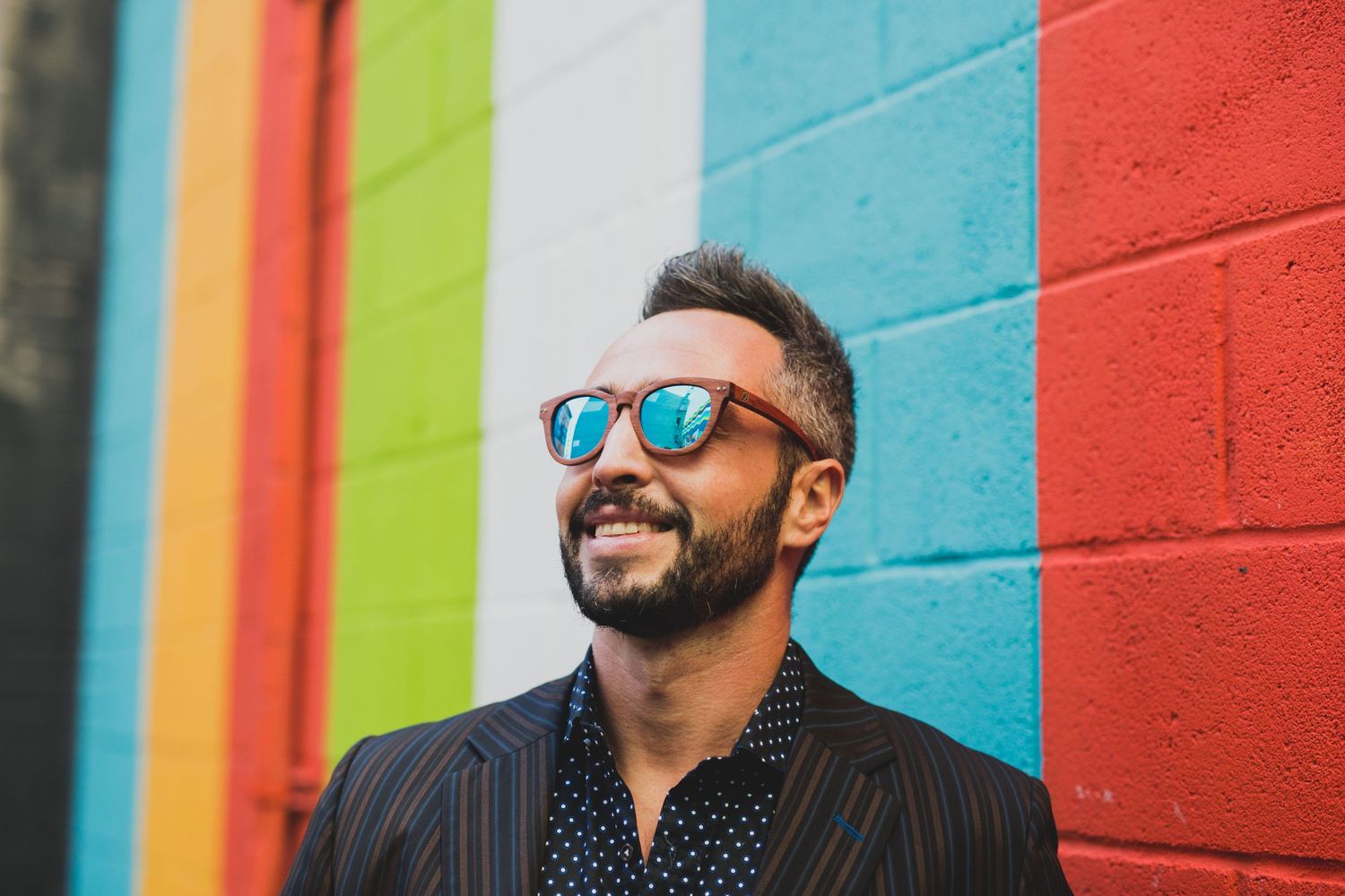 Casual portrait against colorful striped wall wearing polka dot shirt and sunglasses.