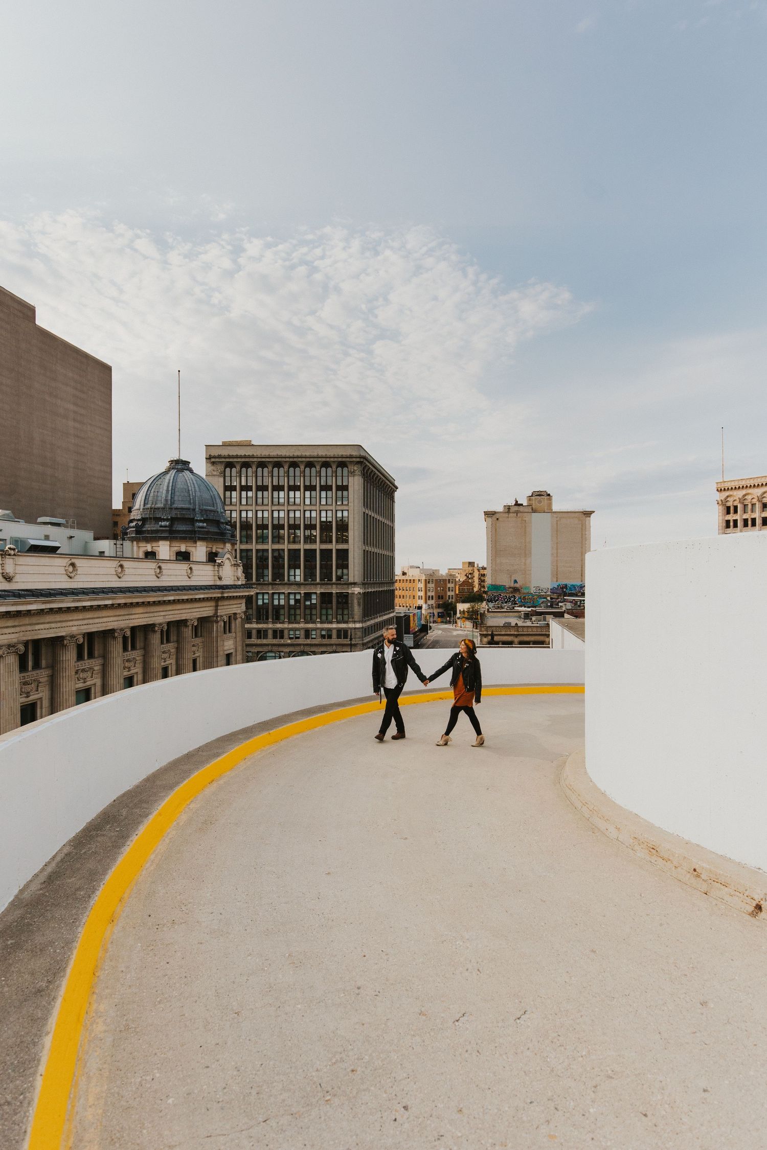 Two people walk hand in hand along a curved yellow line on an urban rooftop.