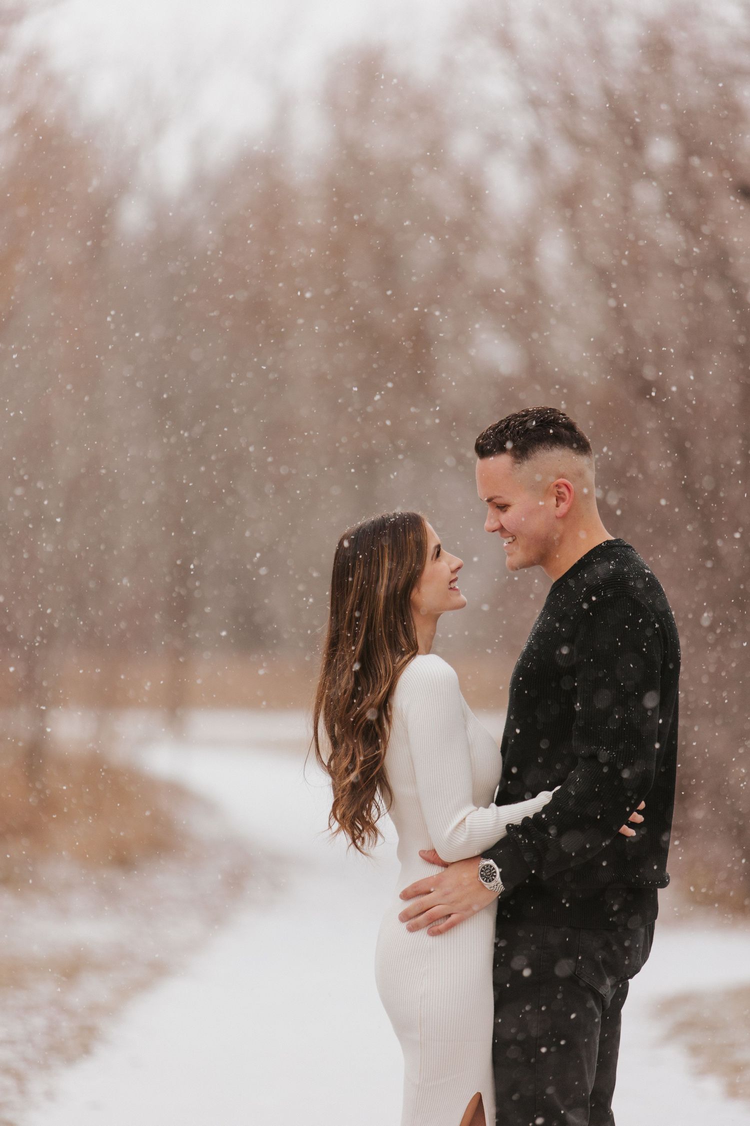 A romantic couple embraces in a snowy winter forest setting during a gentle snowfall.