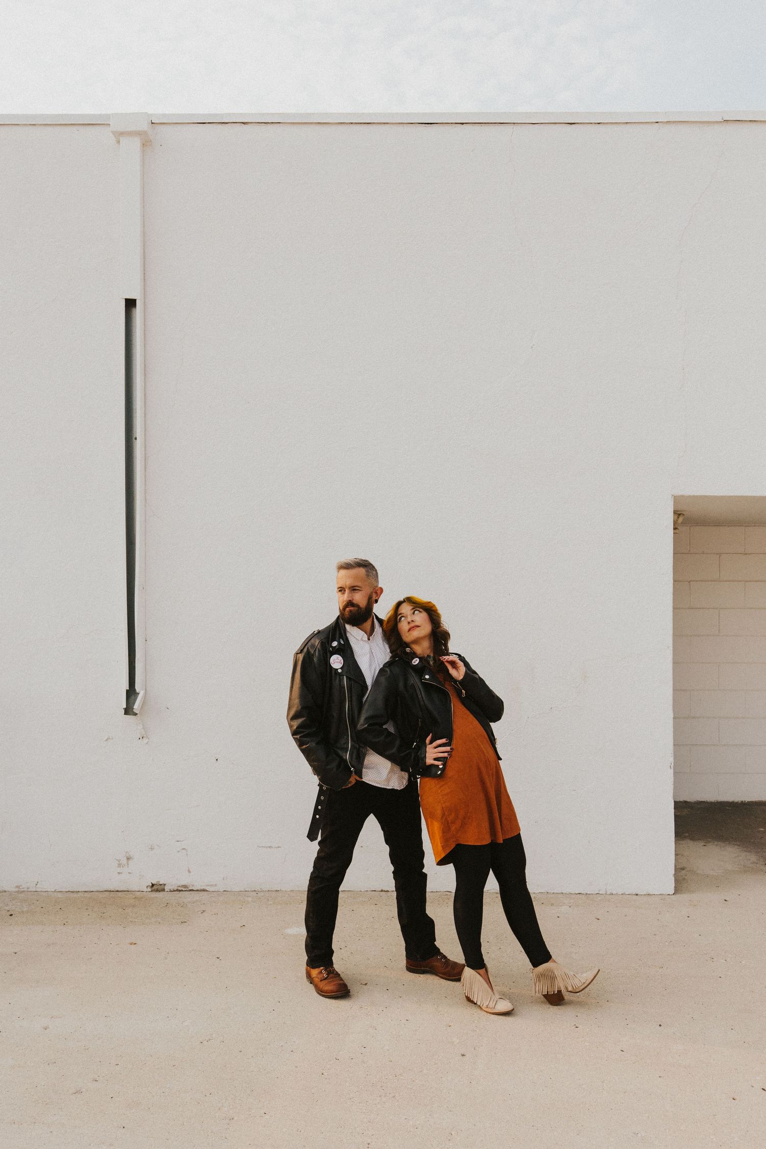 Couple posing against white wall in urban setting wearing black leather and rust-colored clothing.