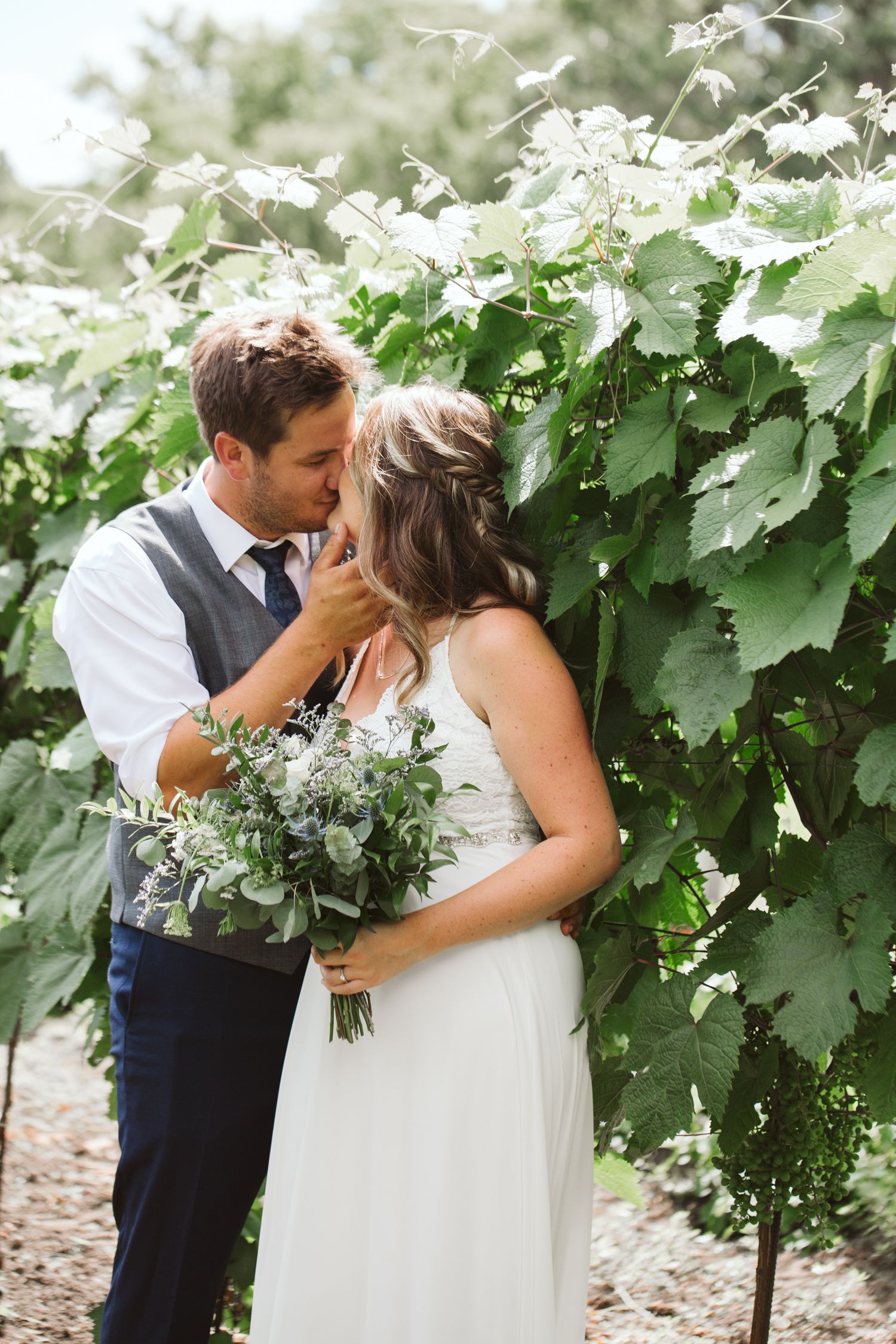Wedding couple embraces near lush grape vines in a vineyard setting.