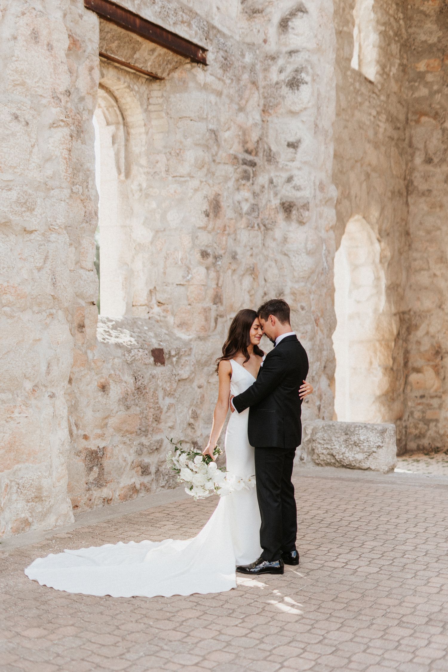 Romantic wedding couple poses against textured stone wall in elegant attire.