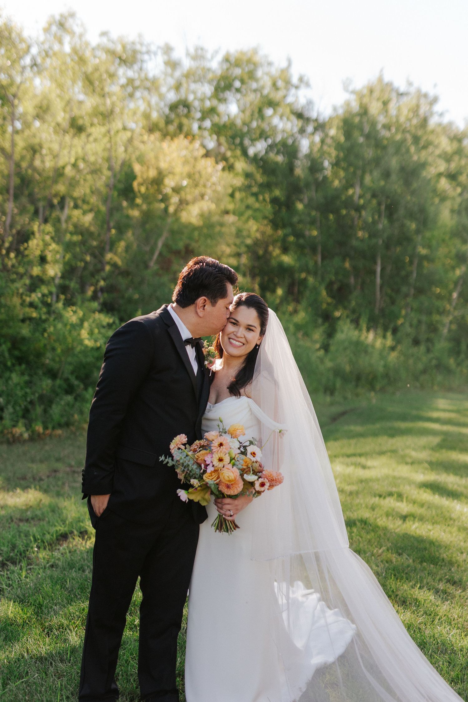 A romantic wedding couple embraces in a sunlit garden setting with flowing white veil and autumn floral bouquet.