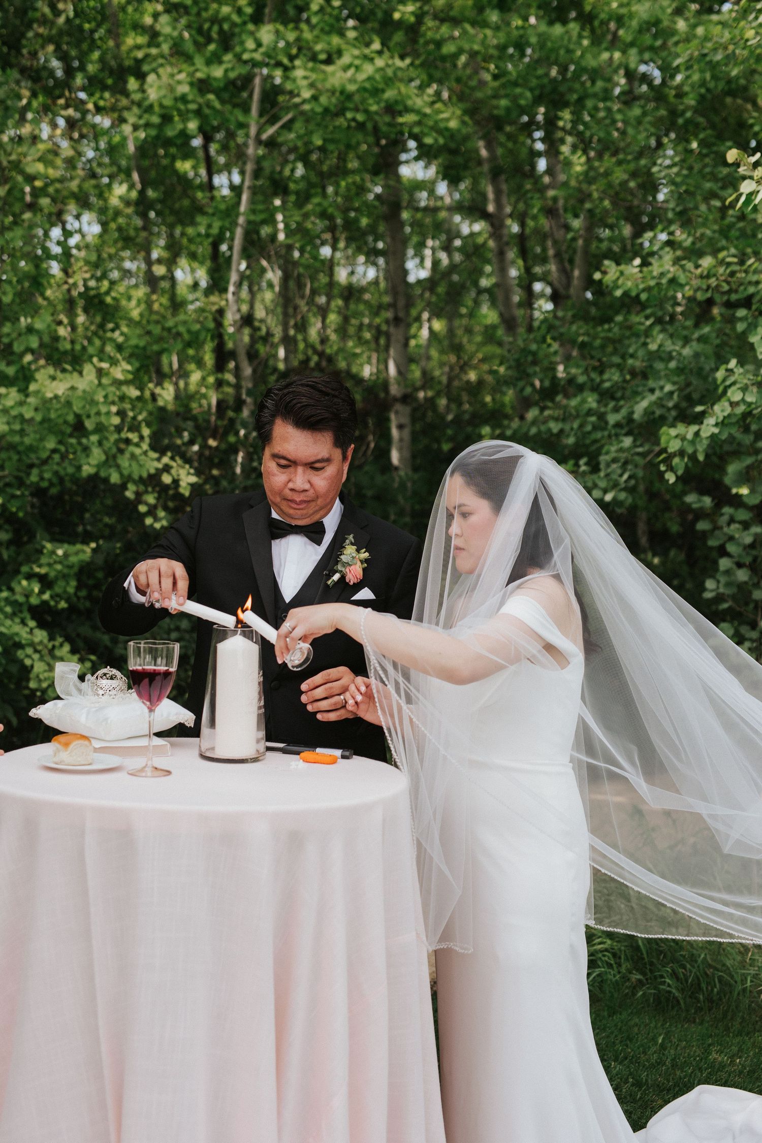 A bride and groom sit at an elegant white-clothed table during their wedding reception outdoors.