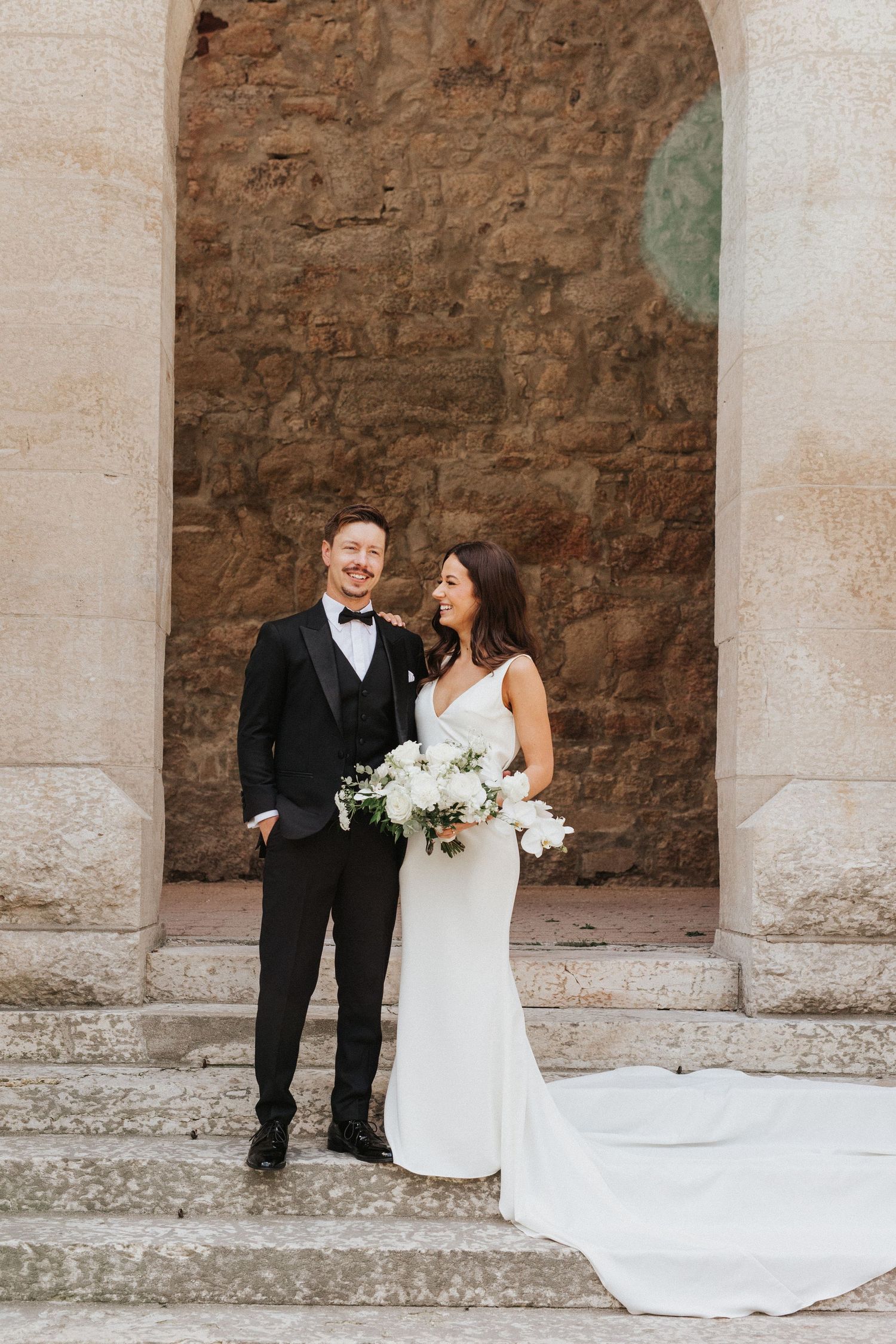 Elegant wedding couple embracing under stone archway with flowing white dress train.