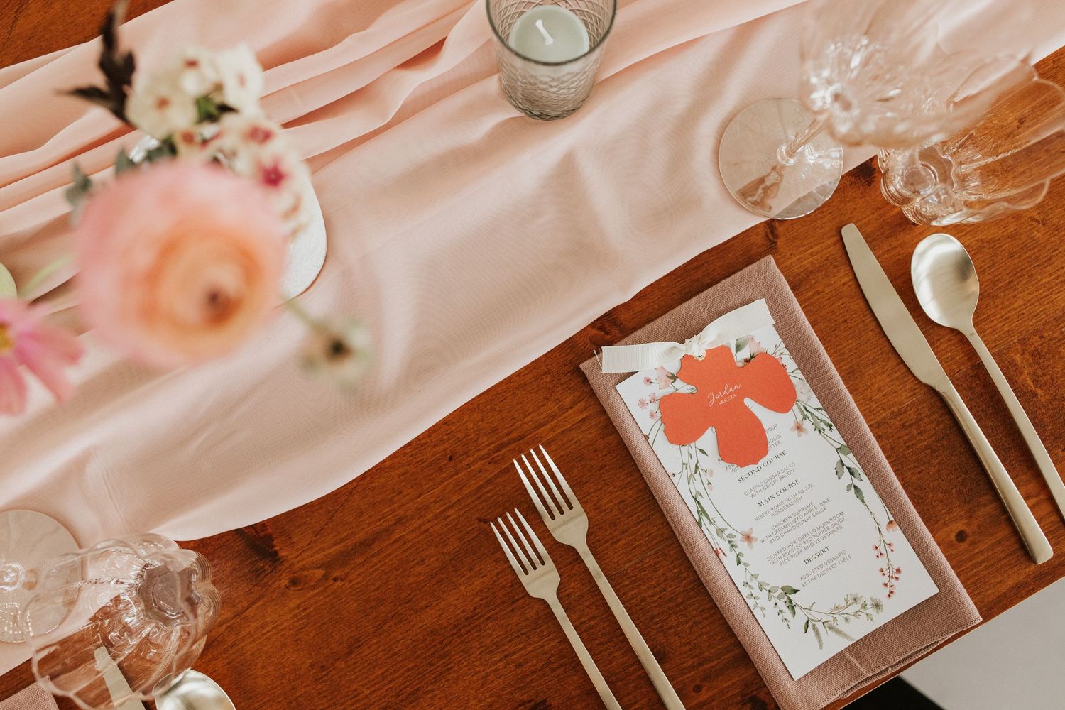Elegant wedding table setting with pink linens, crystal glasses and silver flatware on wooden surface.