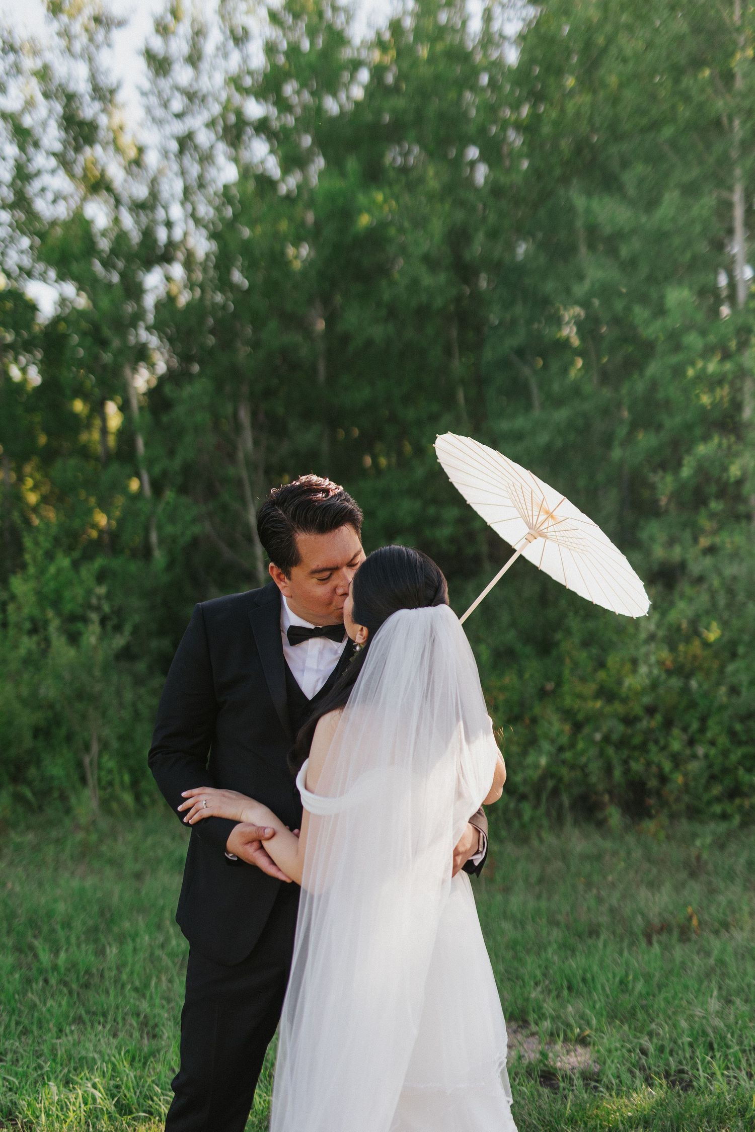 A romantic wedding couple shares an intimate moment under a white parasol in a lush green outdoor setting.