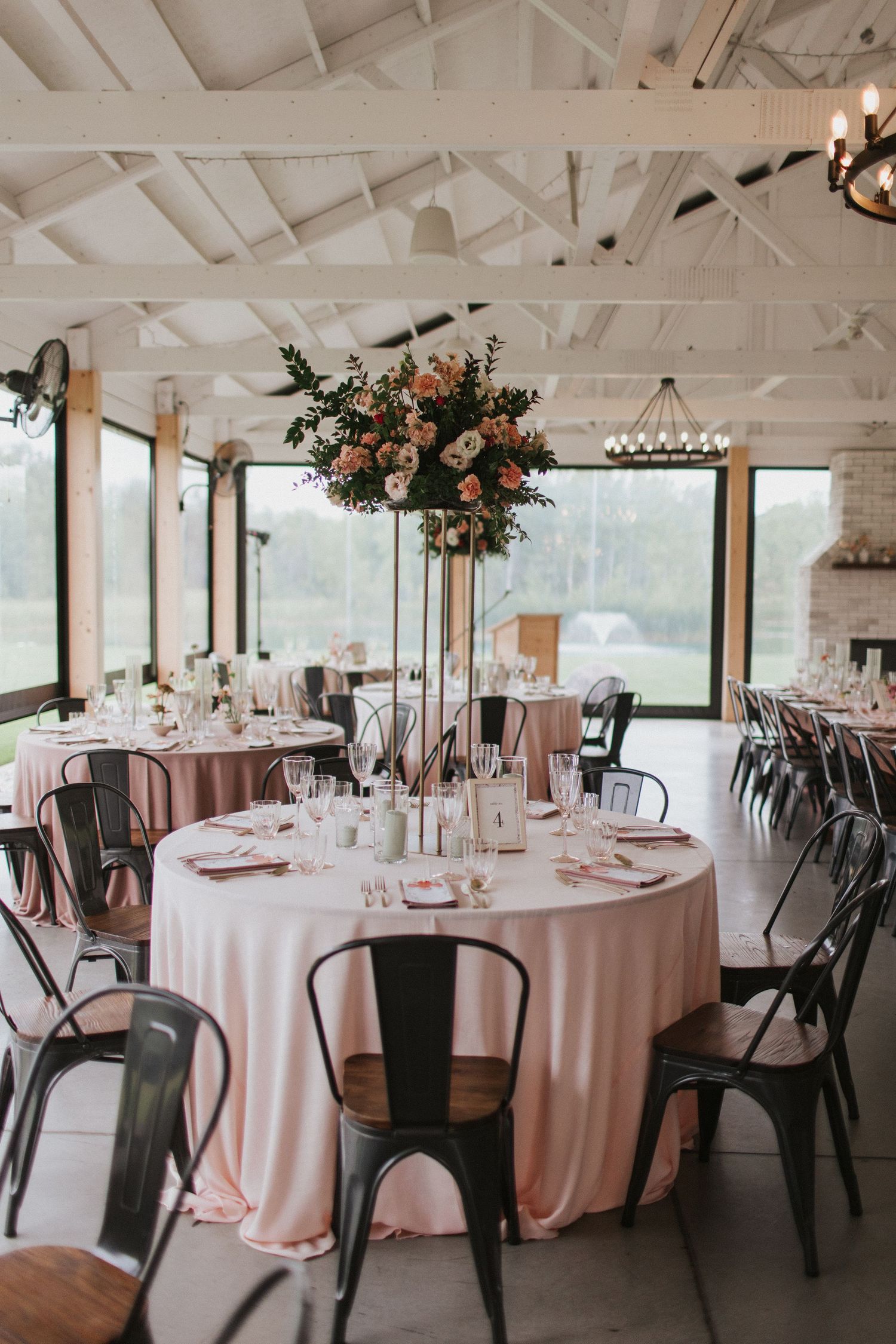 Wedding reception venue with white walls and ceiling featuring round tables with pink linens and black metal chairs.