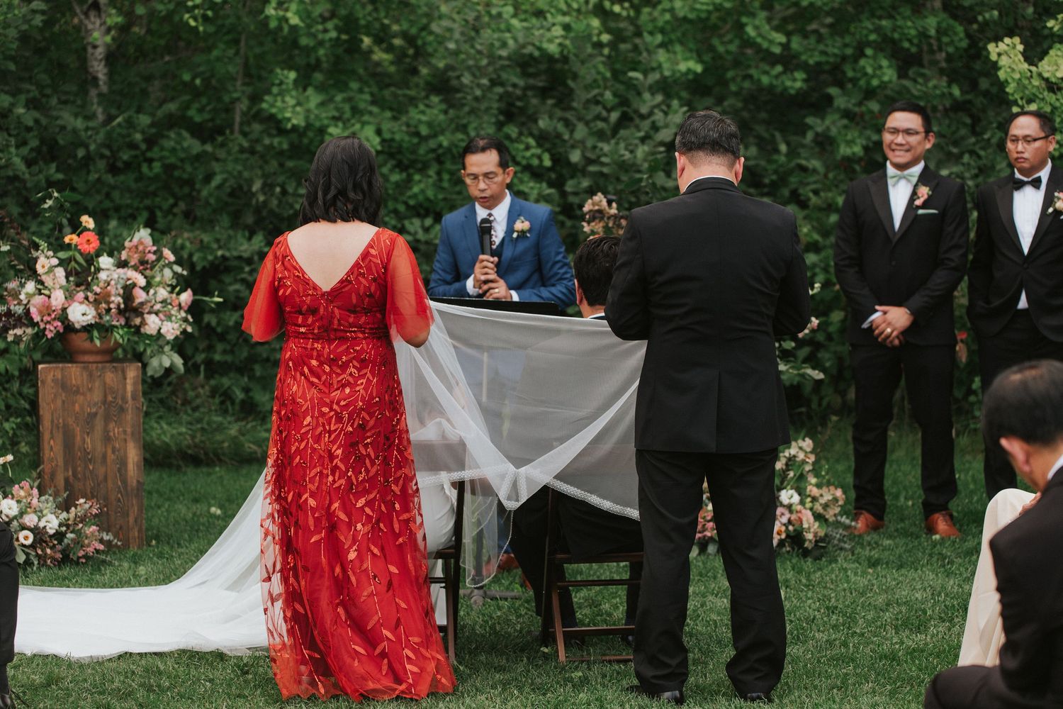 A wedding party poses outdoors in formal attire with bridesmaids in red dresses and groomsmen in black suits.