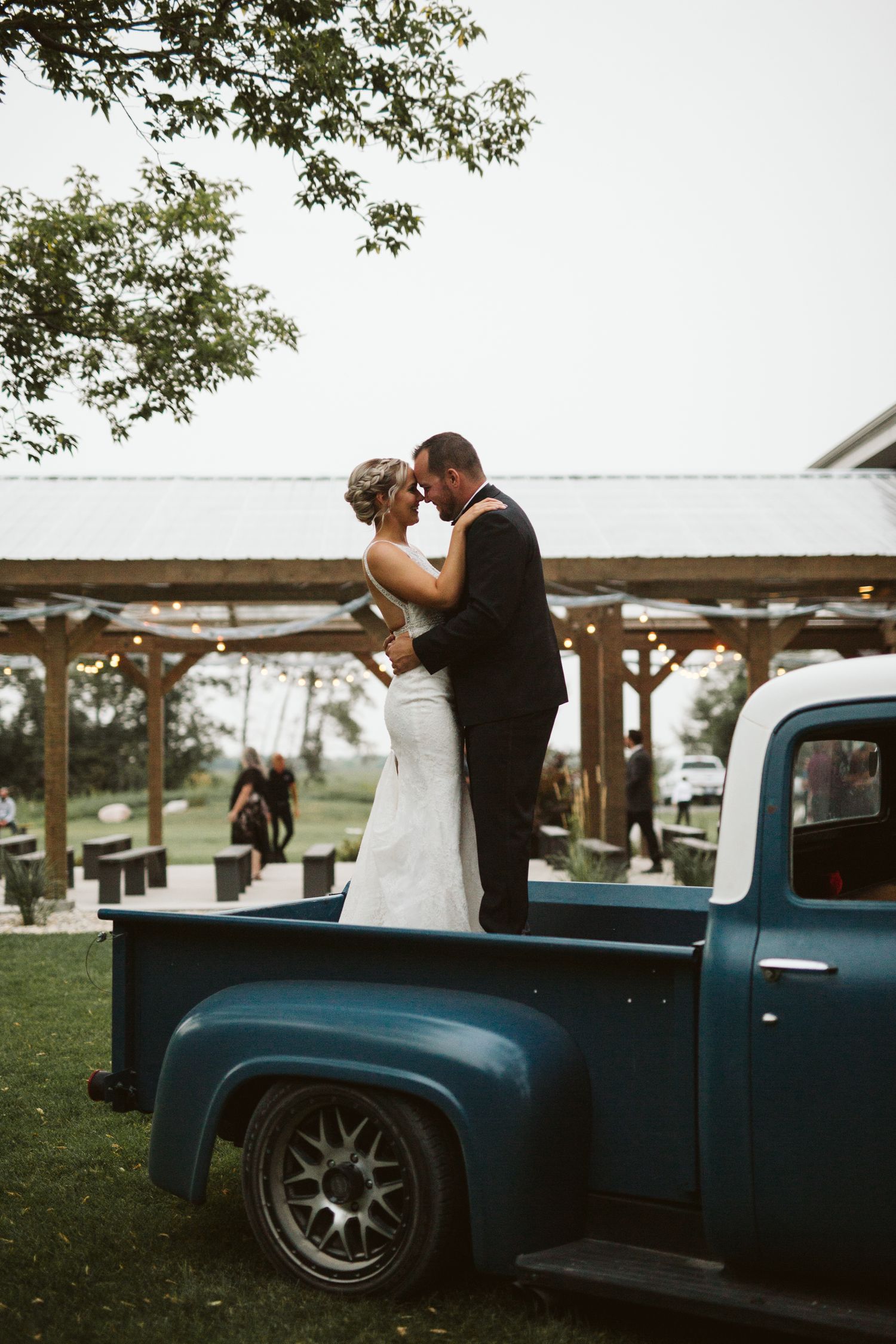 Wedding couple embraces near vintage blue pickup truck outdoors.