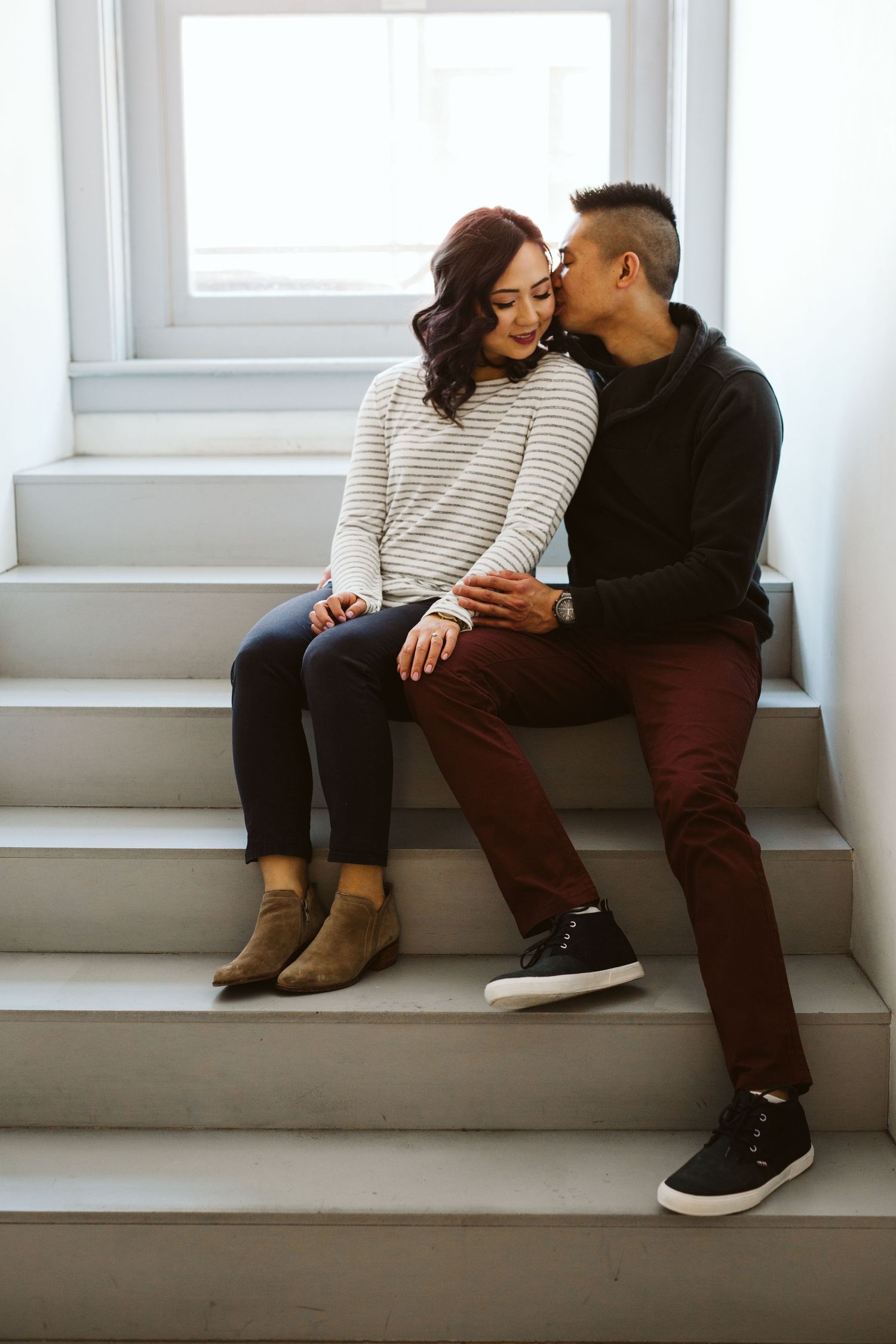 A cozy indoor photoshoot of a couple sitting on white steps by a window.