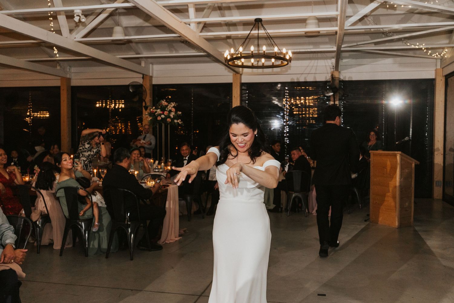Elegant wedding reception venue with exposed beams, chandeliers and guests mingling on dance floor.