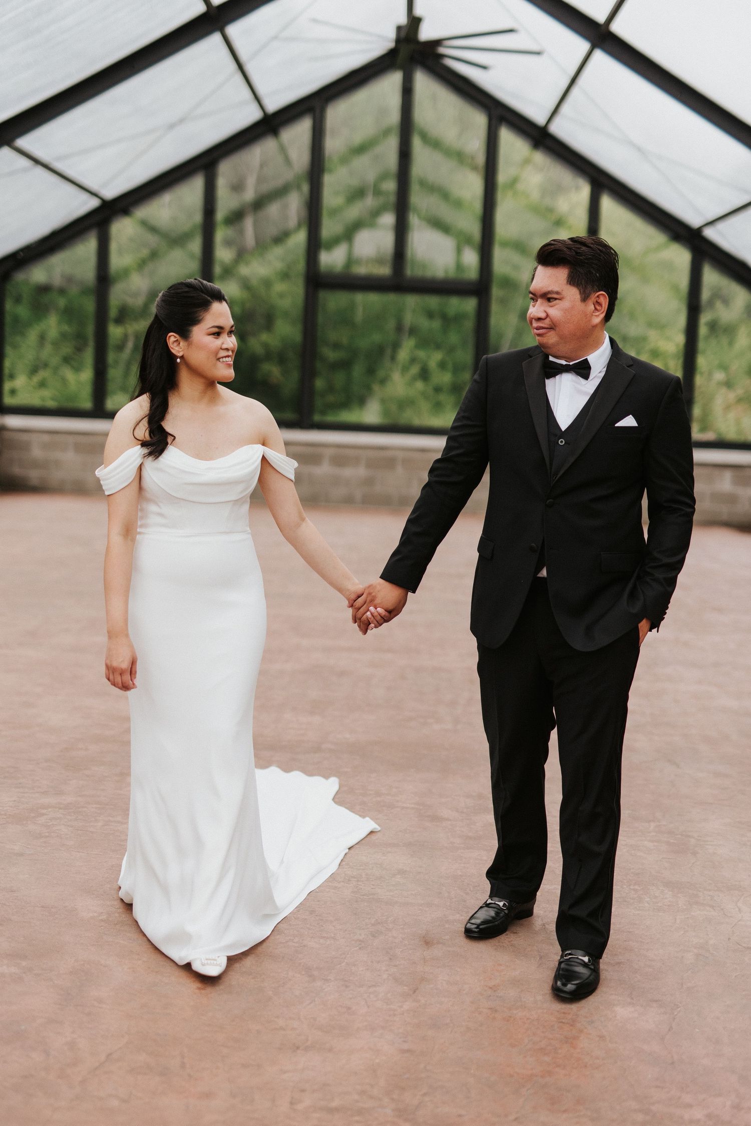 Newlyweds share a romantic moment walking together in a greenhouse setting.