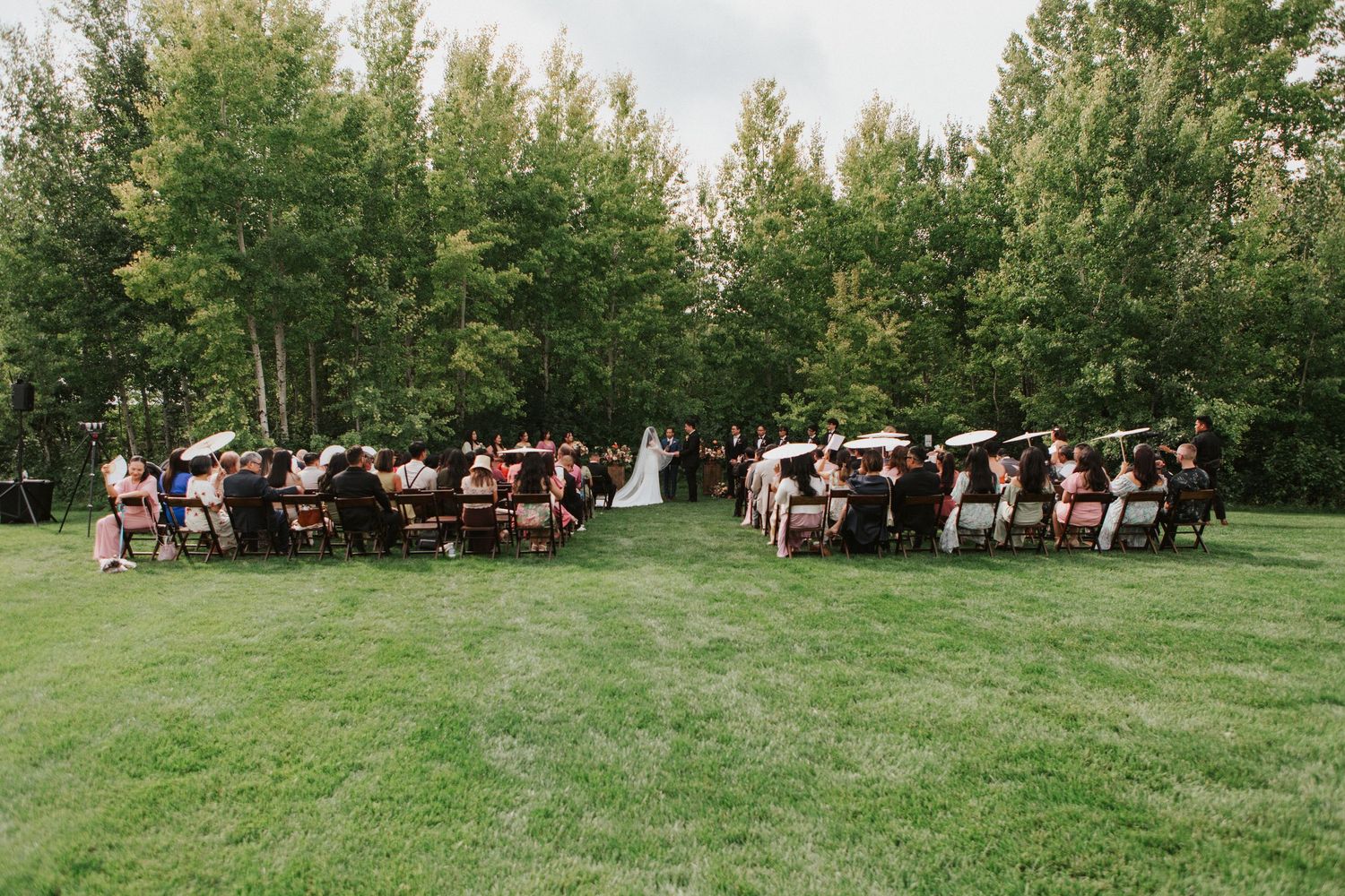 An outdoor wedding ceremony setup with guests seated on white chairs surrounded by green trees.