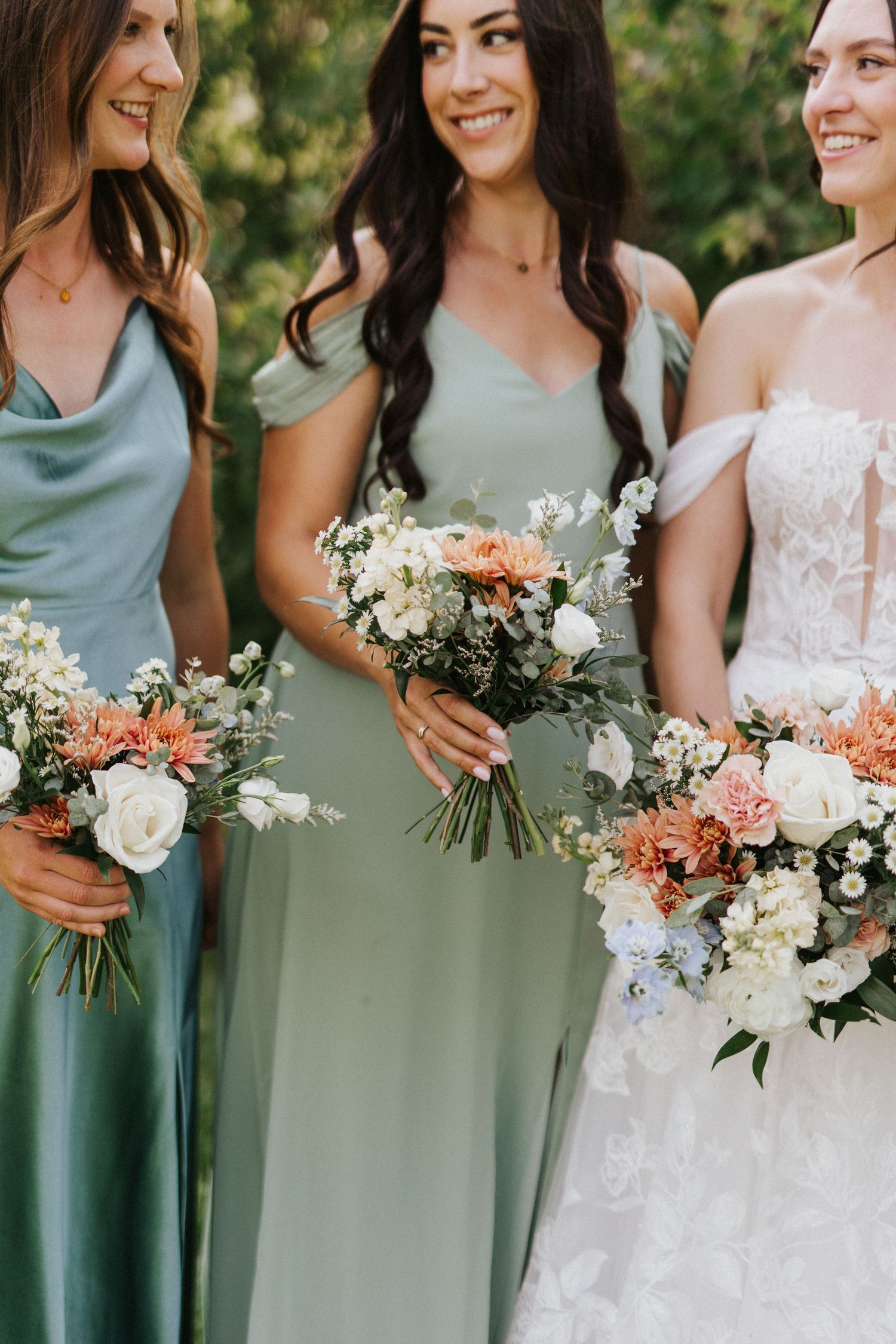 Bridesmaids in sage green dresses hold bouquets while standing together.