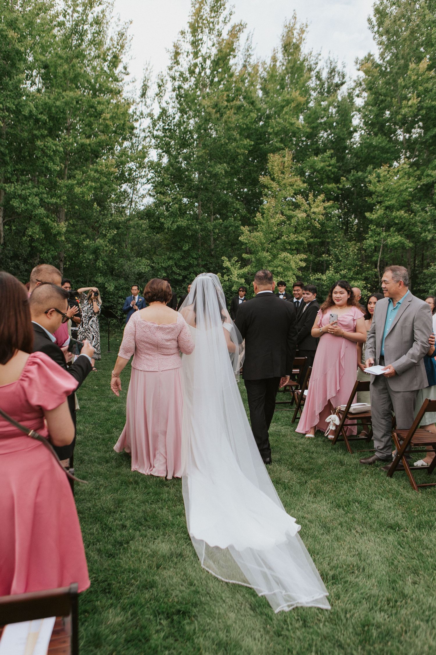 Wedding guests in pink dresses gather on a lawn as a bride in a flowing veil walks through the outdoor ceremony.