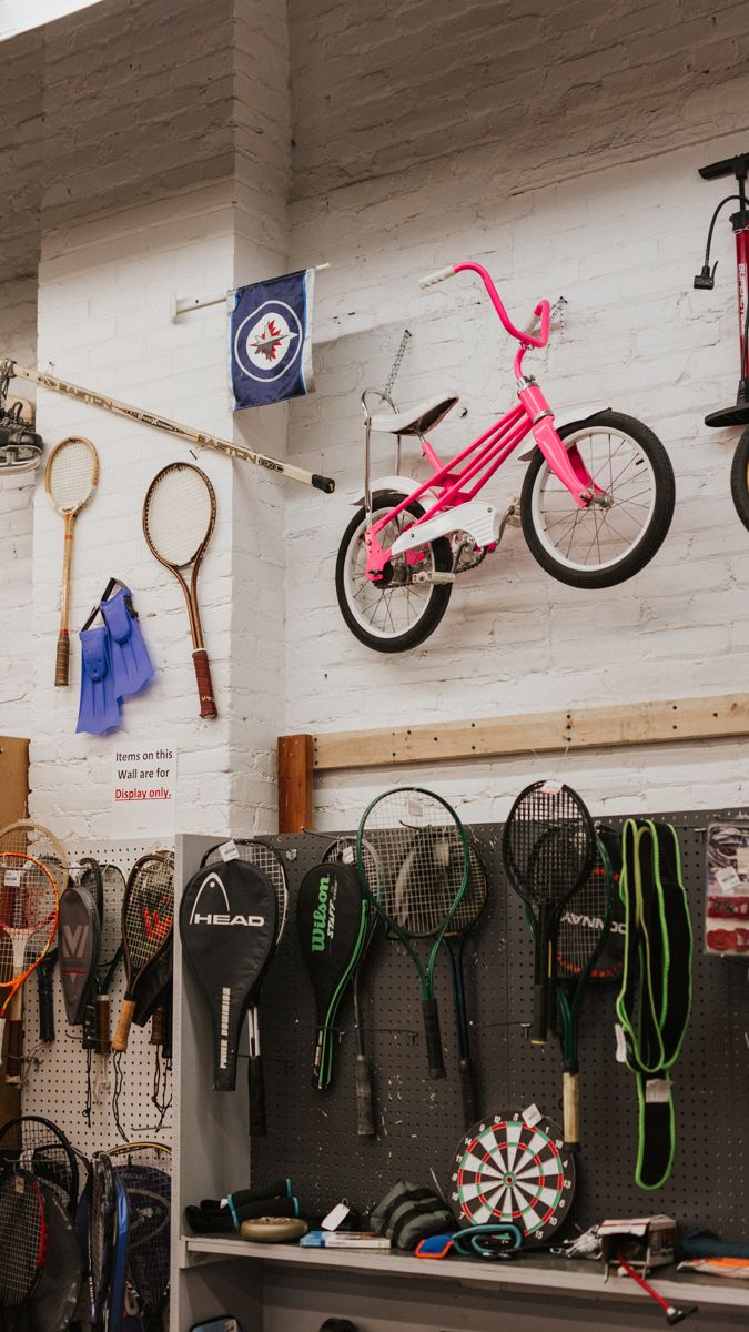 Sports equipment and accessories displayed on white wall with dartboards and pink bicycles hanging above.